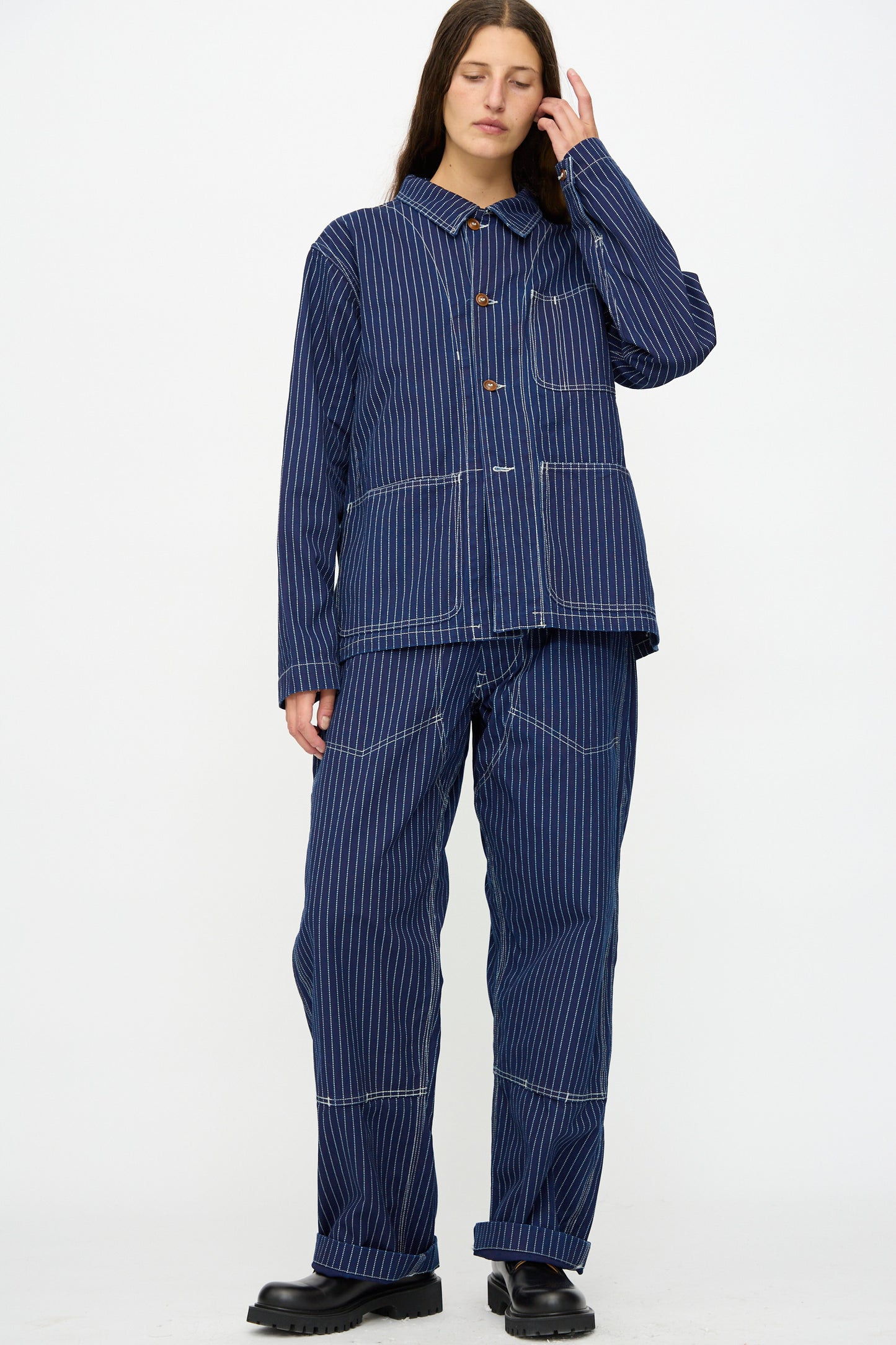 A person stands against a white background, wearing the Henri Pant in Wabash by As Ever with a matching blue pinstripe jacket and black shoes, touching their hair with one hand.