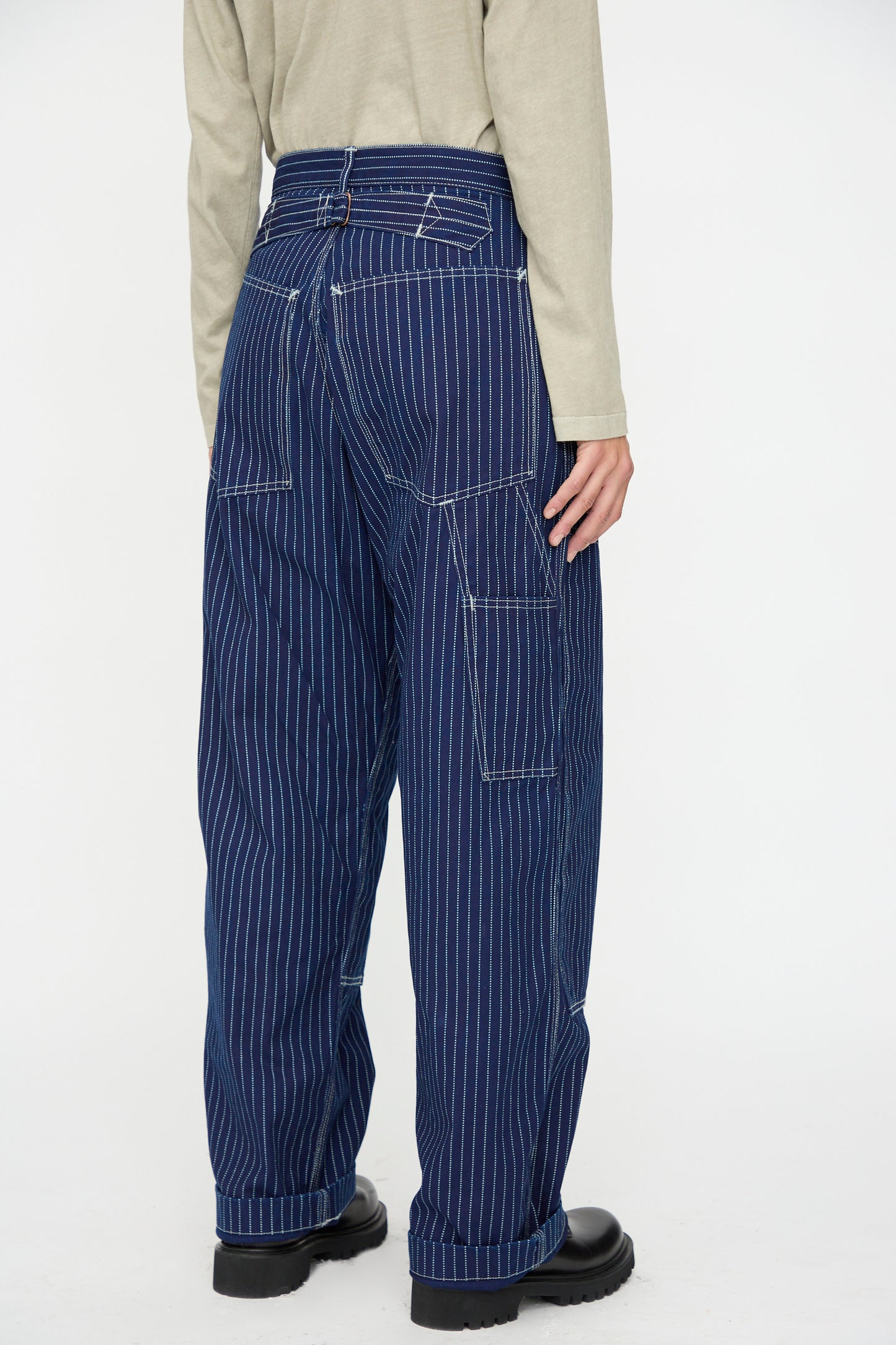 Person standing with their back to the camera, wearing As Ever's Henri Pant in Wabash—blue pinstriped, oversized, multi-pocket work pants—paired with a beige long-sleeve shirt and black shoes against a plain background.