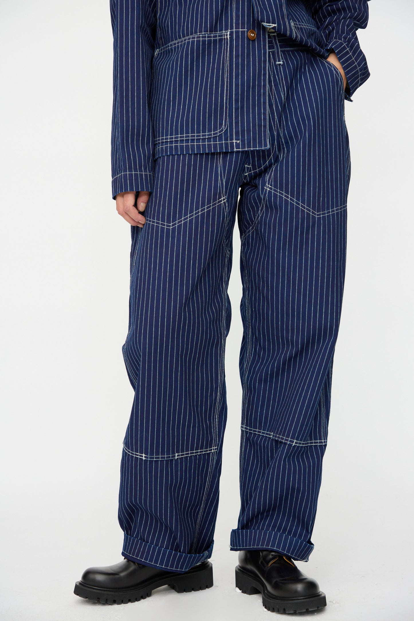 A person wears As Ever's Henri Pant in Wabash, styled as an oversized workwear suit with a matching jacket and black shoes, standing against a plain white background.