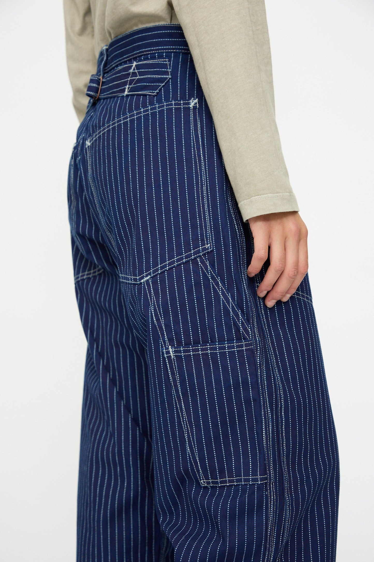A person shown from the waist down at a side angle wears As Ever's Henri Pant in Wabash, which features loose-fitting, blue pinstriped vintage military styling with visible stitching, paired with a beige long-sleeve shirt.