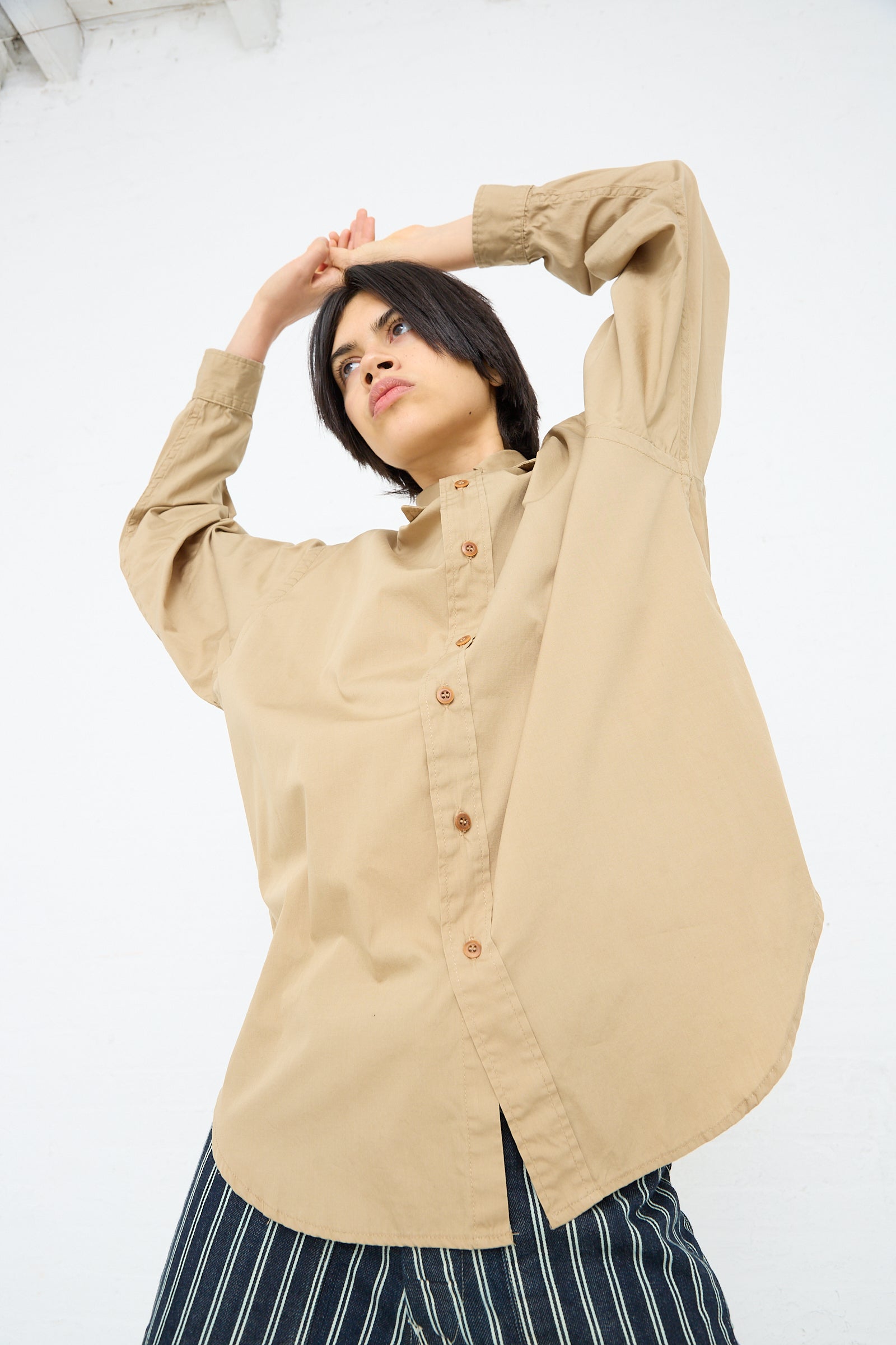 A person in an oversized Largo Shirt in 40's Khaki by As Ever and striped trousers posing against a white background with one arm raised and hand on head.