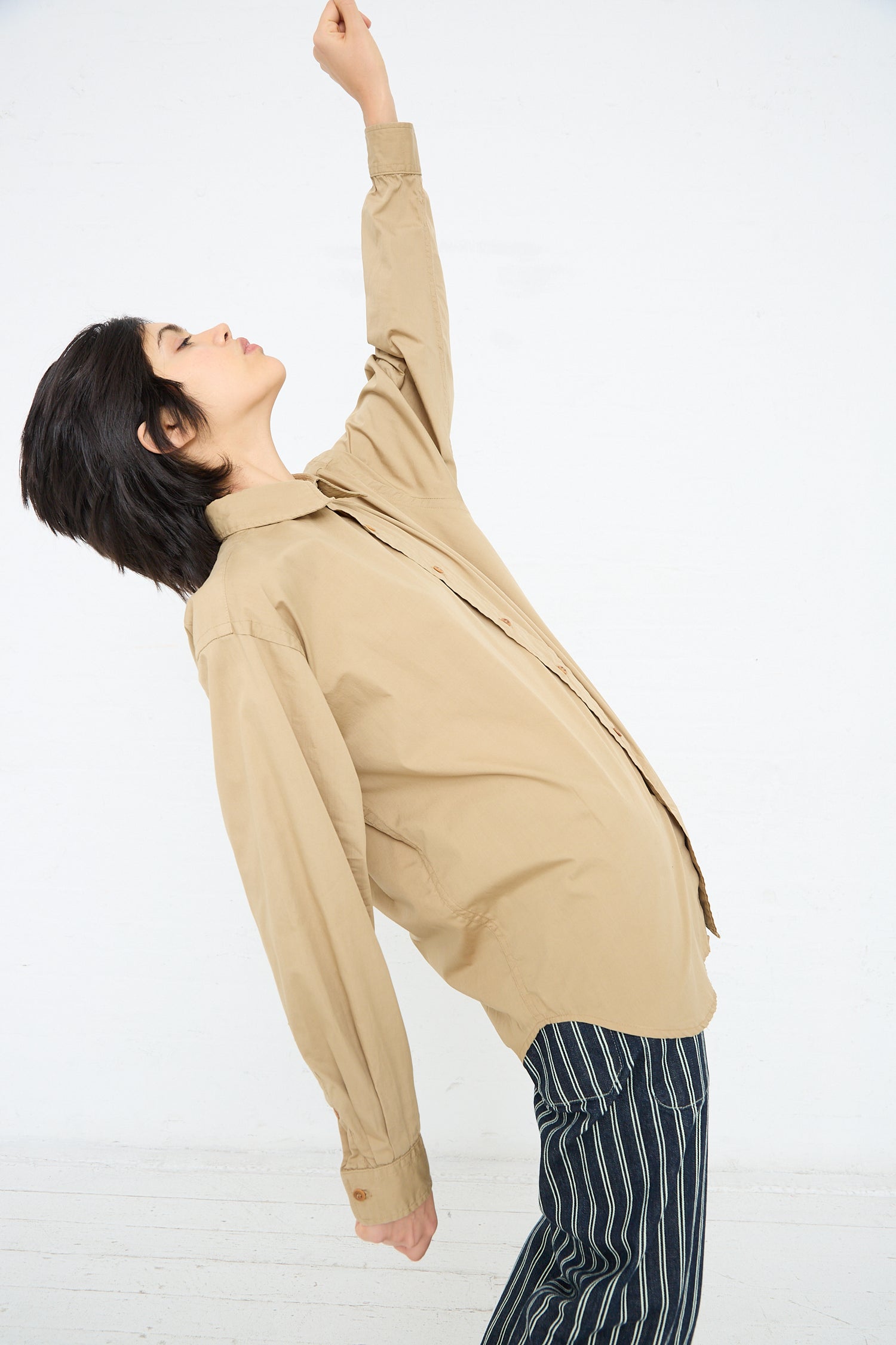 A person in a beige jacket and striped pants stretching one arm upwards while standing against a white background, donning an As Ever Largo Shirt in 40's Khaki.