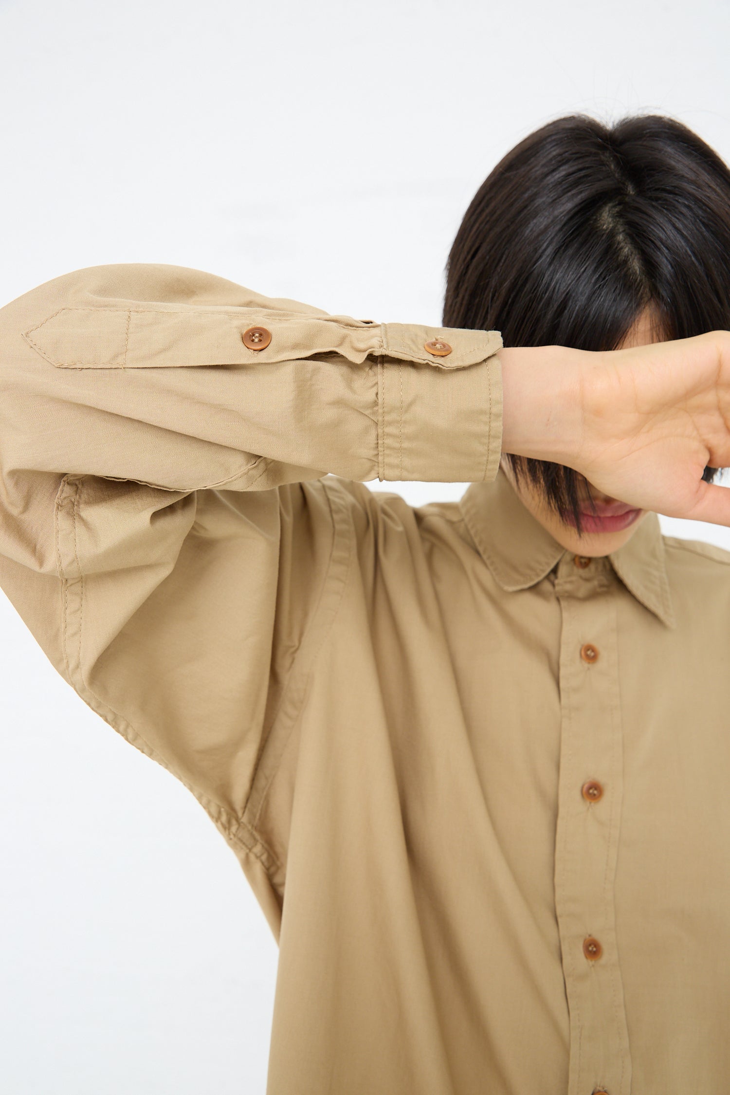 A man in an oversized, tan As Ever Largo Shirt in 40's Khaki, made of 100% cotton, covering his face with his hands.