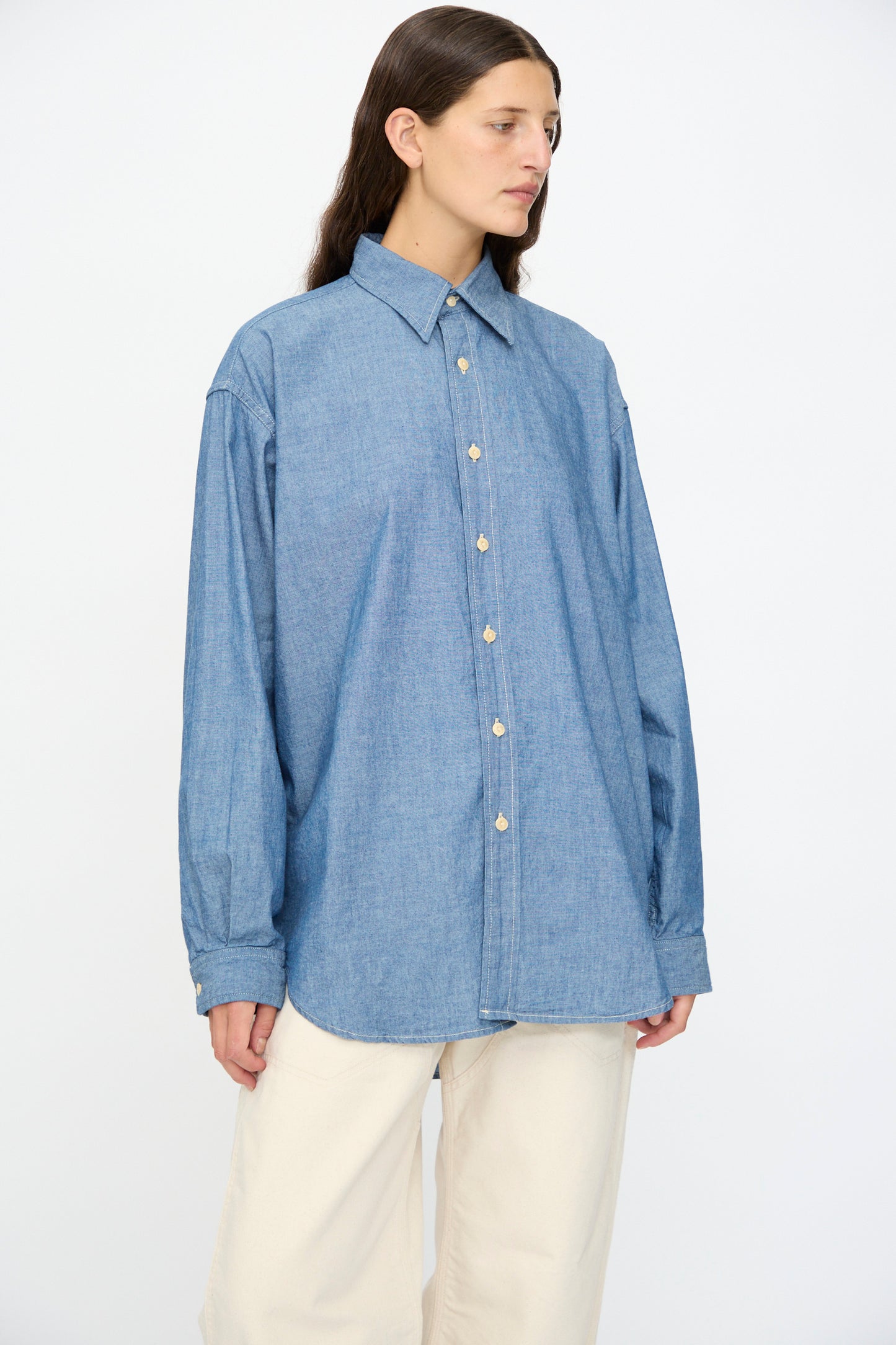 Model wears the As Ever Largo Shirt in Chambray, styled with off-white pants, standing against a plain white background.