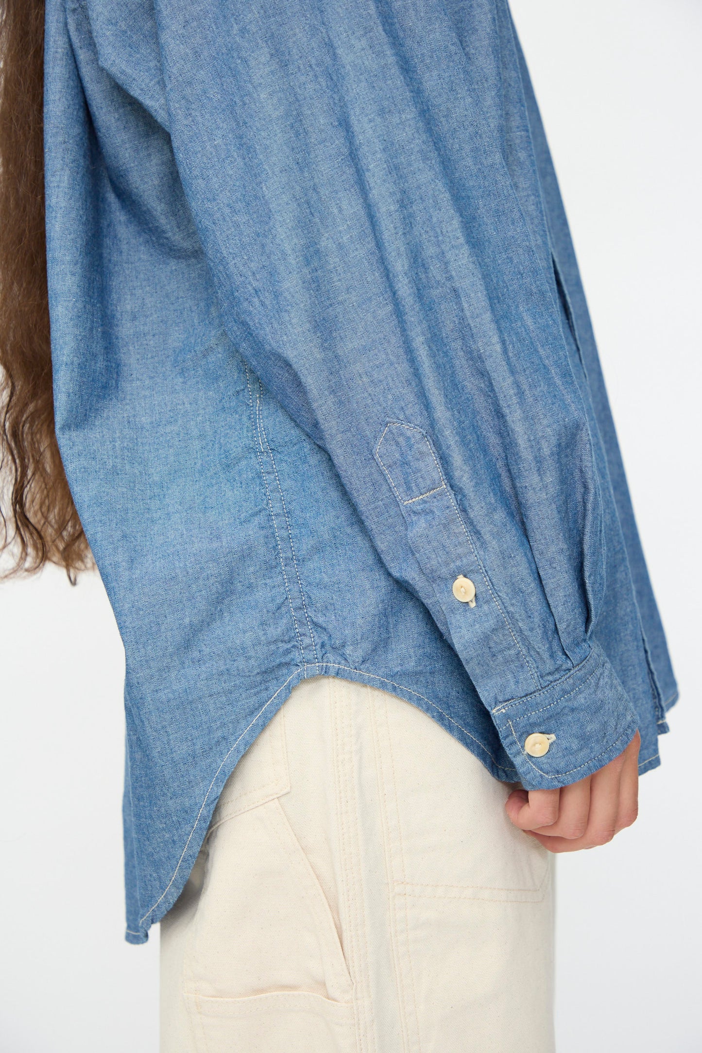 A person wears the As Ever Largo Shirt in Chambray with off-white pants, shown from the side with one arm relaxed and hair visible.