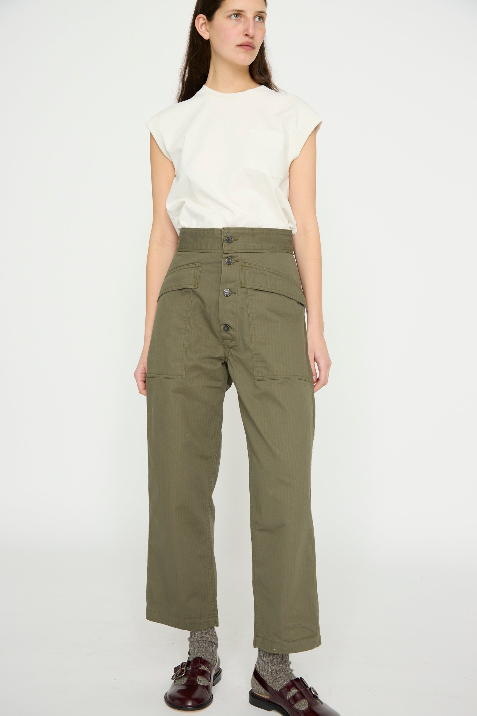 Wearing As Ever's Tanker in Olive, a person stands against a plain background, showcasing the high-waisted pants with brown shoes.