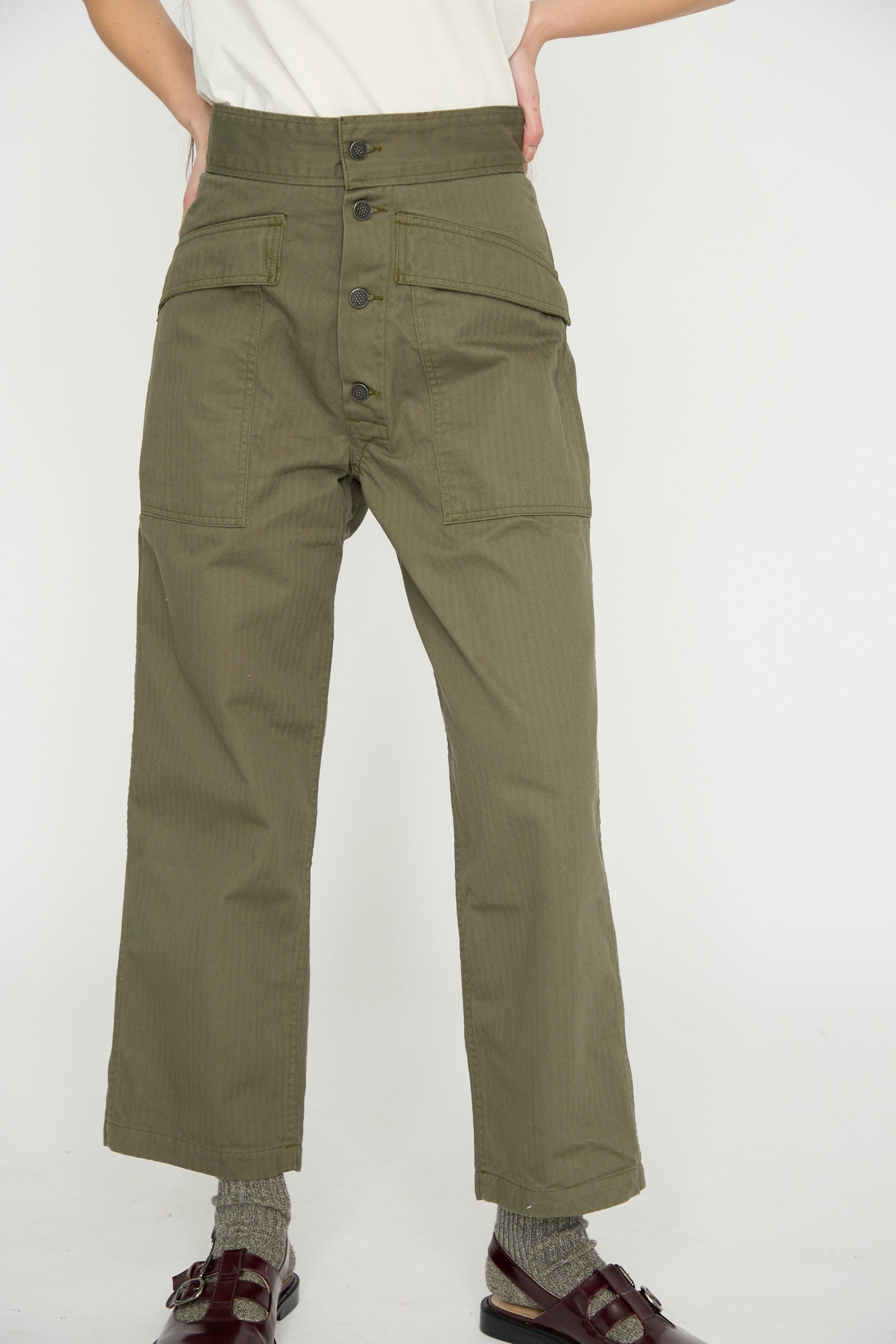 Wearing As Ever's "Tanker in Olive" high-waisted herringbone twill pants with buttons and pockets, a person stands against a plain background, paired with a white shirt, gray socks, and brown sandals.