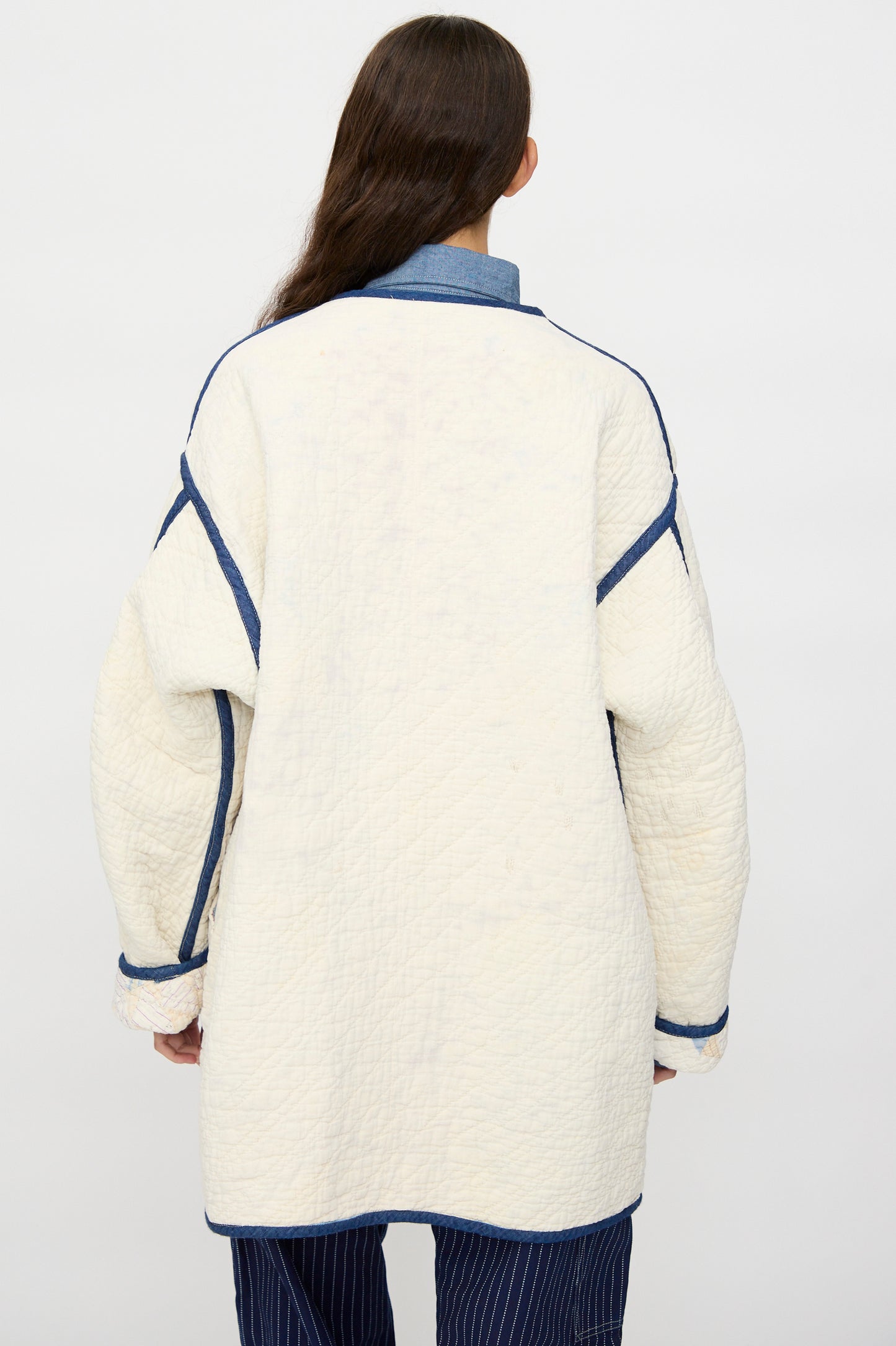 A person with long brown hair stands with their back to the camera wearing As Ever’s Vintage Quilt Coat in Red and Blue, paired with a blue collared shirt and pinstriped pants.