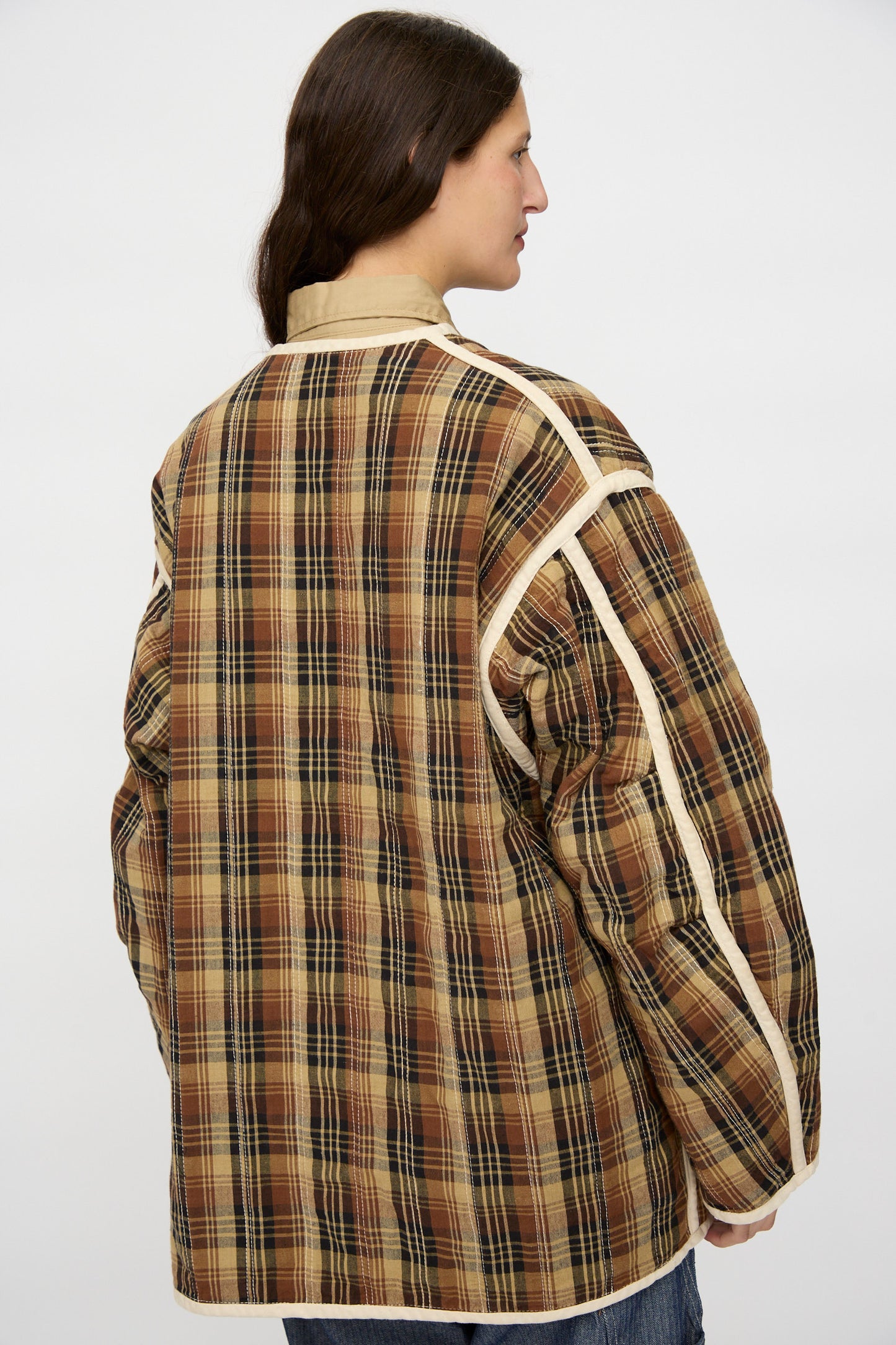 Person with long brown hair wearing the As Ever Quilt Coat in HBT Natural, a brown and beige plaid quilted jacket with white trim, shown from the back against a plain background.