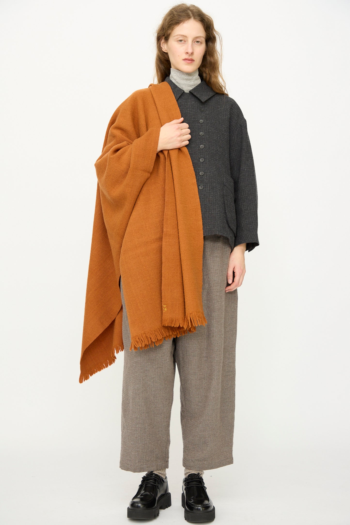 A person stands against a plain background, wearing gray clothes and black shoes, with the Auntie Oti Wool Throw in Rust, featuring a fringe edge, draped over one shoulder.
