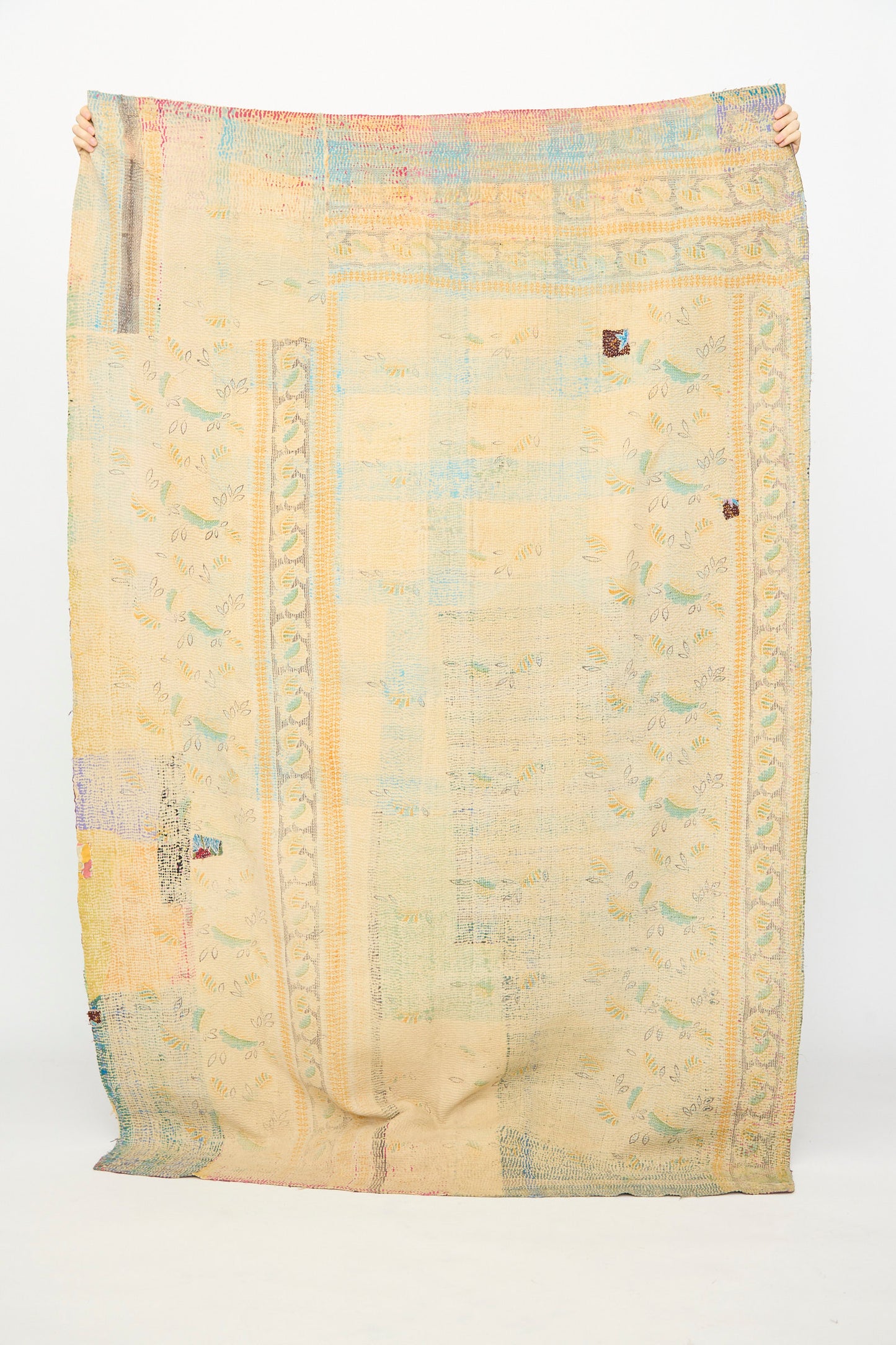 A person holds up the One of a Kind Vintage Kantha Quilt III by Auntie Oti, a large beige quilt with pastel patterns and colorful patches, handcrafted from upcycled cotton, shown against a plain white background.