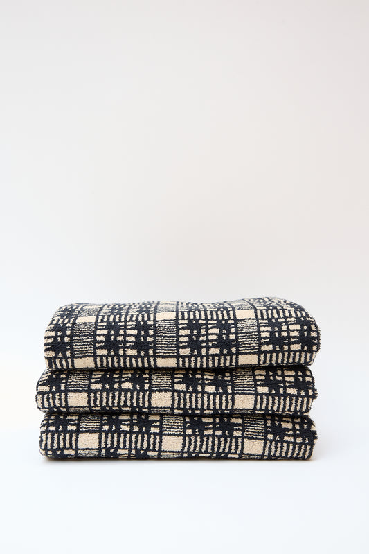 Three folded Alma Bath Towels with a black and white geometric pattern, made in Portugal, stacked on each other against a light gray background.