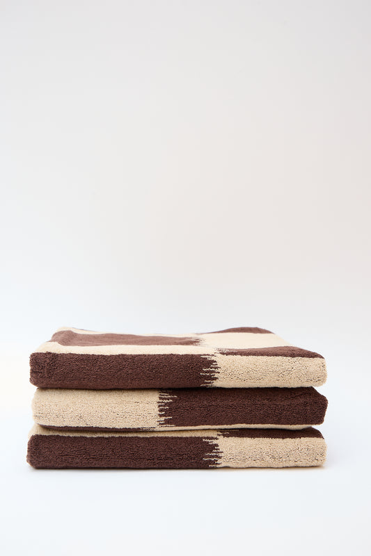 A stack of three Autumn Sonata Karin Bath Towels in brown and beige with Ikat weaving technique patterns, set against a plain light background.