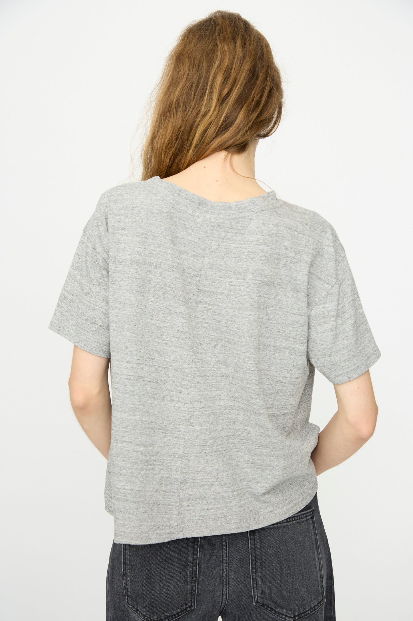 A person with long, light brown hair stands facing away in a relaxed fit B Sides Atlas Tee in Heather Grey and dark gray jeans, set against a plain white background.