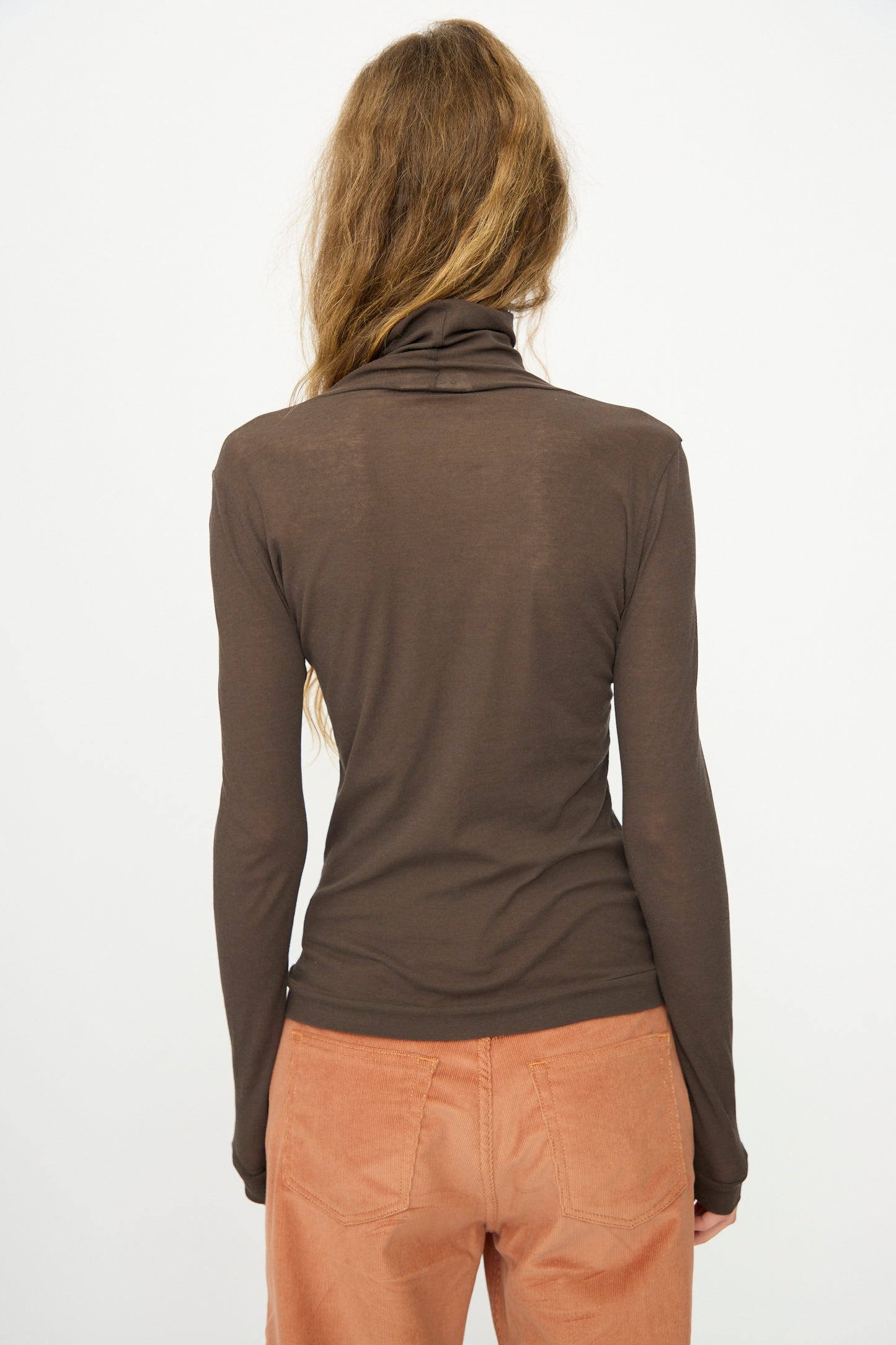 Person with long, wavy hair stands with their back to the camera against a plain background, wearing B Sides' Jana Turtleneck in Olive Drab and light brown pants.