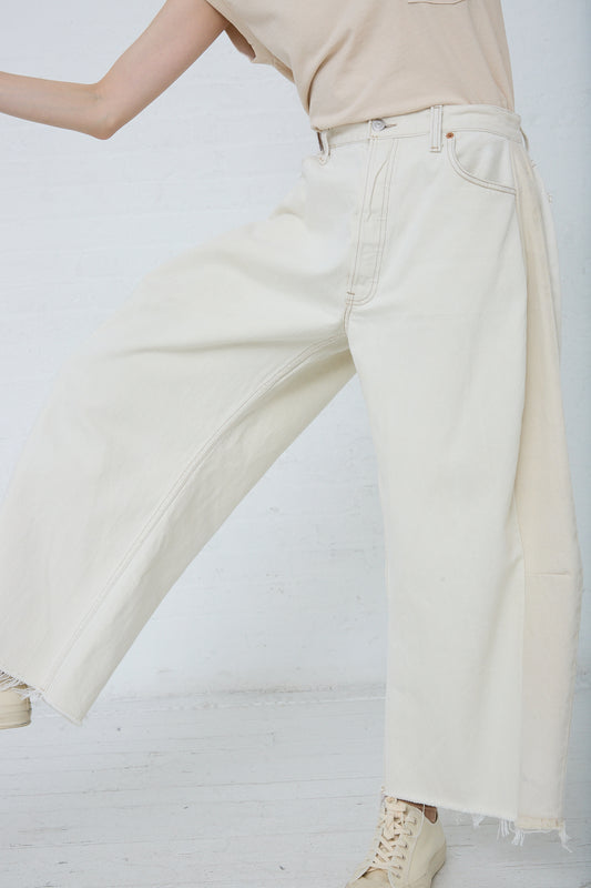 A person standing against a white wall wearing B Sides' Lasso Jean in Vintage White, slouchy fit jeans with frayed hems and beige boots.
