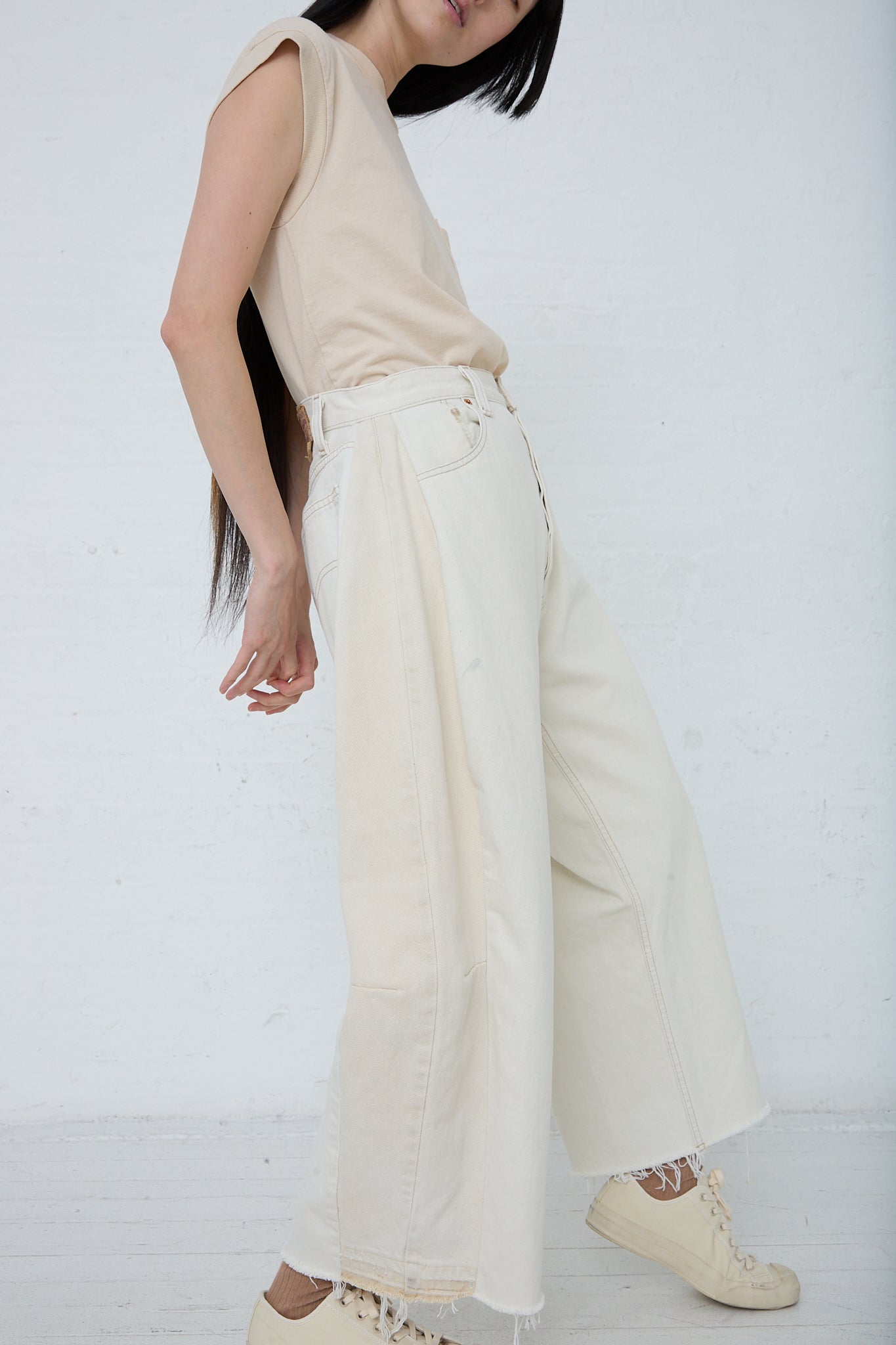 Woman standing against a white background wearing a sleeveless top and wide-leg, reworked denim jeans with frayed hems, paired with beige sneakers by B Sides featuring the Lasso Jean in Vintage White.
