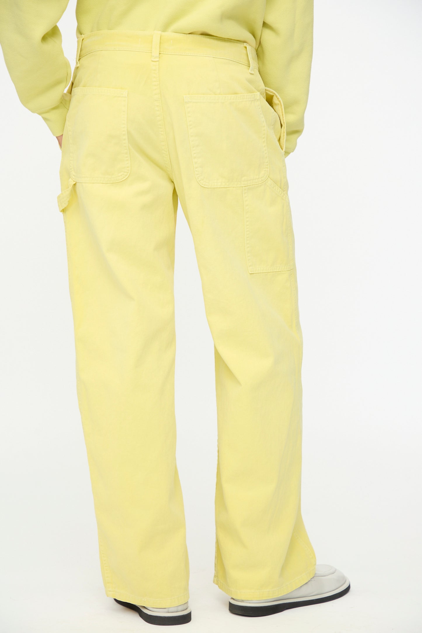 A person is shown from the back against a plain white background, wearing B Sides' Painter Pant in Faded Celery Overdye—loose-fitting, pale yellow cotton chinos with large back pockets—paired with a matching yellow top.