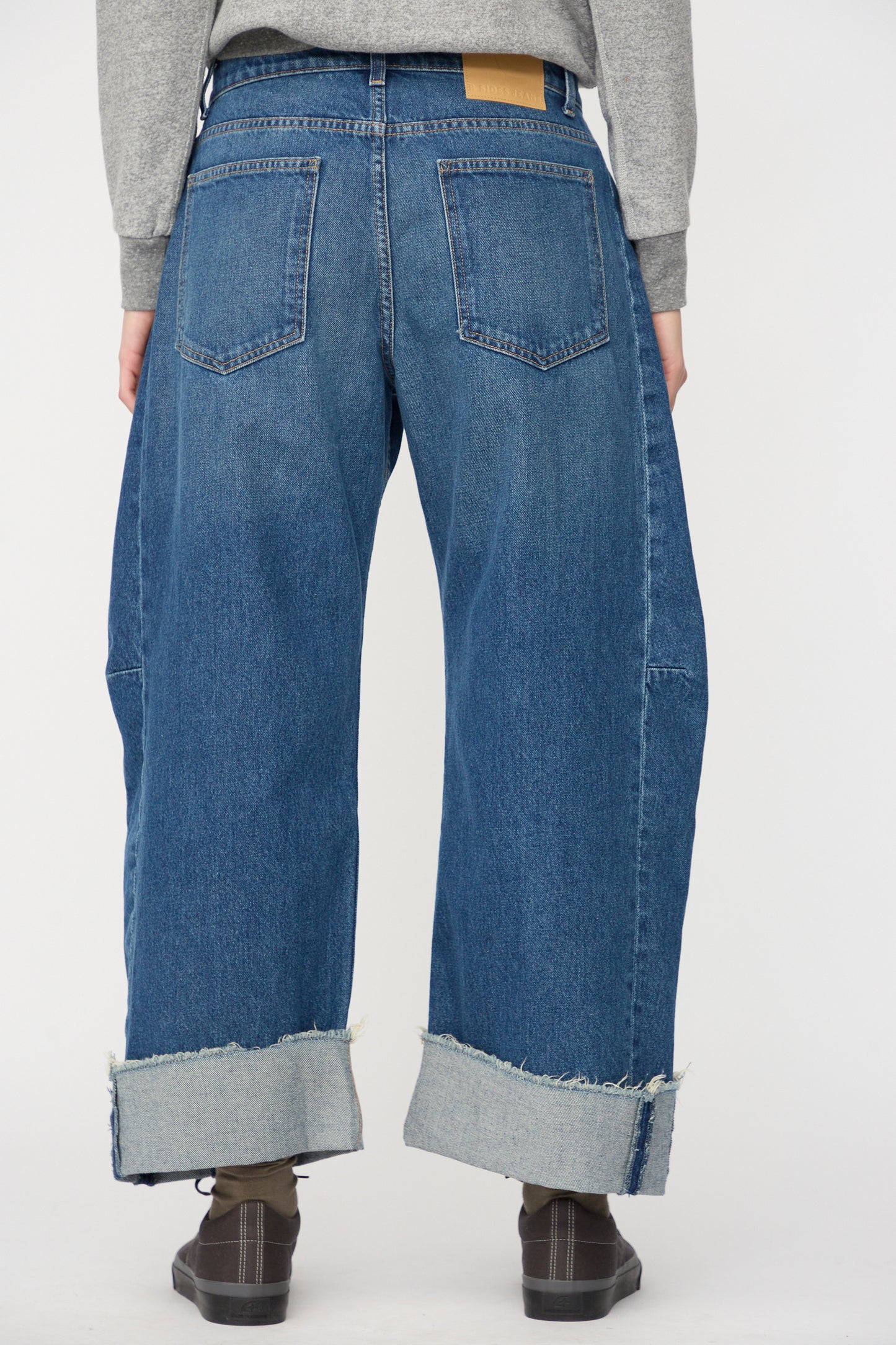 Person wearing B Sides Relaxed Cuffed Lasso Jean in Cate Wash with large cuffed hems, gray socks, and brown shoes, seen from the back against a plain background.