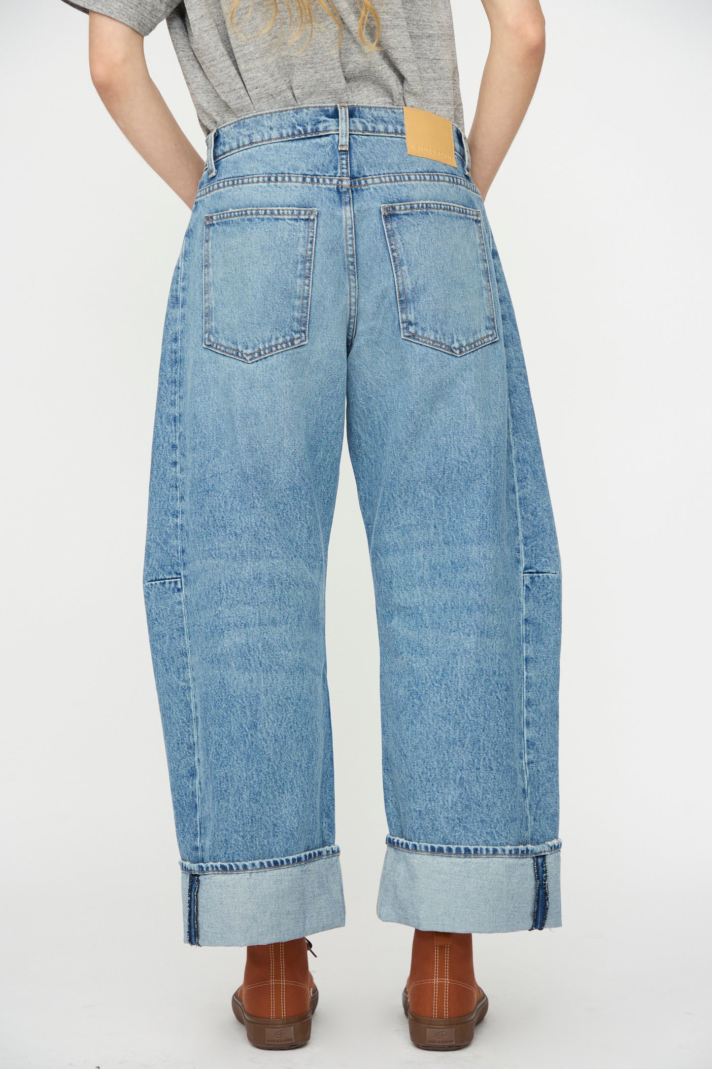 Model wears the B Sides Relaxed Cuffed Lasso Jean in Jane Wash, featuring wide-leg, mid-rise heavy cotton denim with light blue cuffs and brown shoes, shown from the back against a plain background.