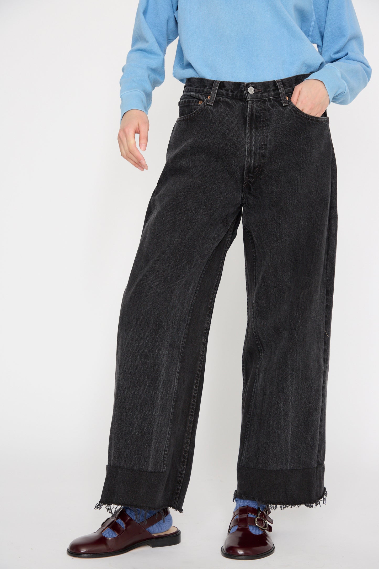 A model wears B Sides' Reworked Culotte in Vintage Black, made from upcycled vintage Levi's 501's, paired with a blue top and burgundy shoes against a plain background.