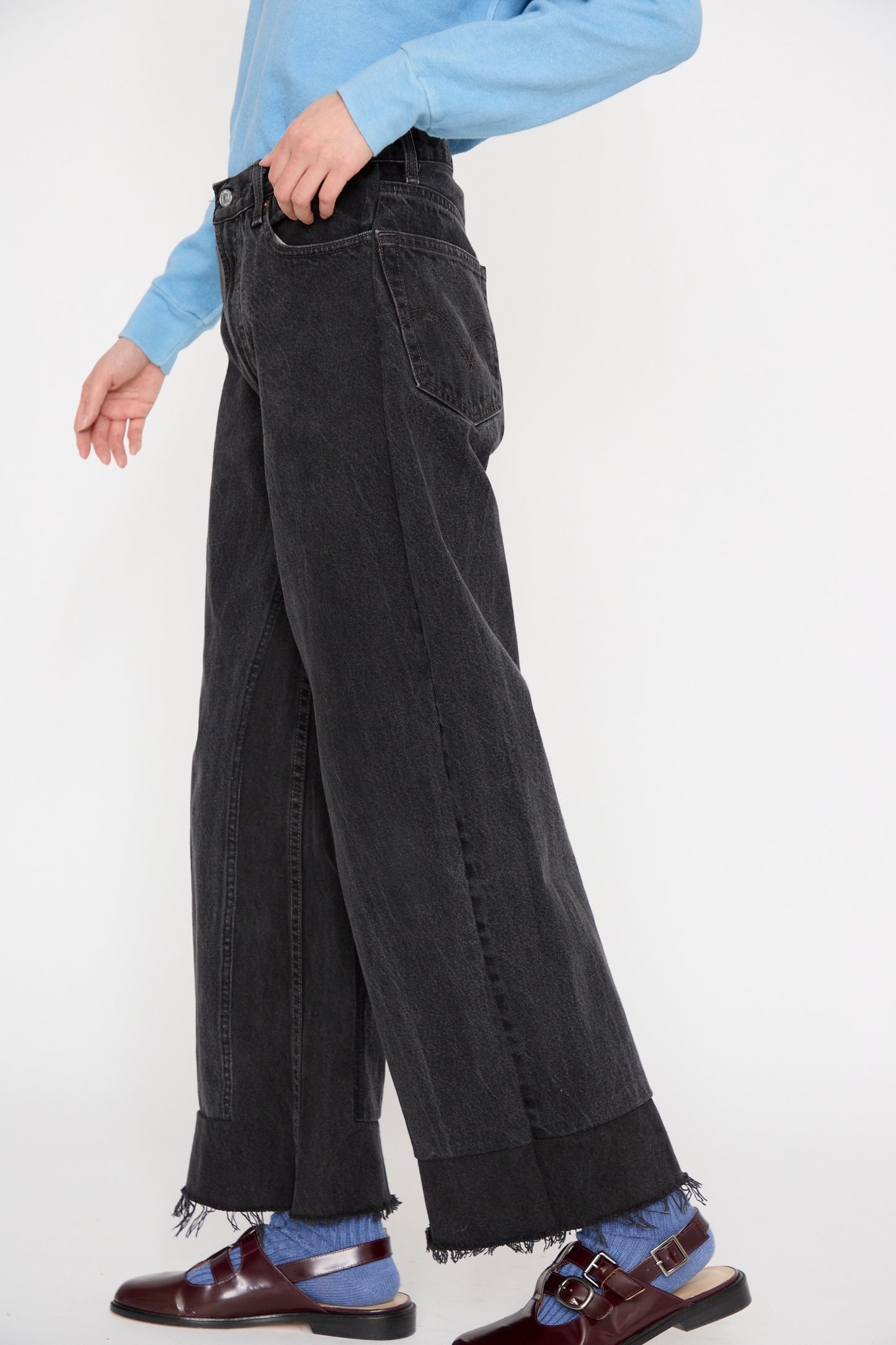 A person in a blue shirt wearing B Sides Reworked Culotte in Vintage Black, complemented by brown shoes and blue socks, walks against a plain white backdrop.