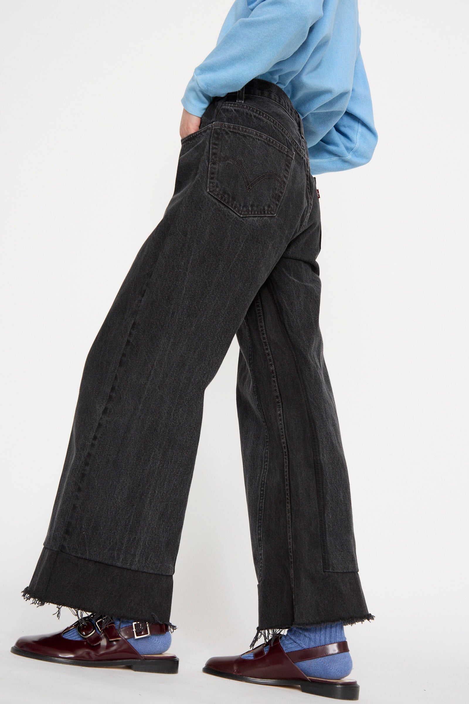 A person in a blue shirt and B Sides Reworked Culotte in Vintage Black, with frayed hems, paired with blue socks and maroon loafers, stands against a white background.