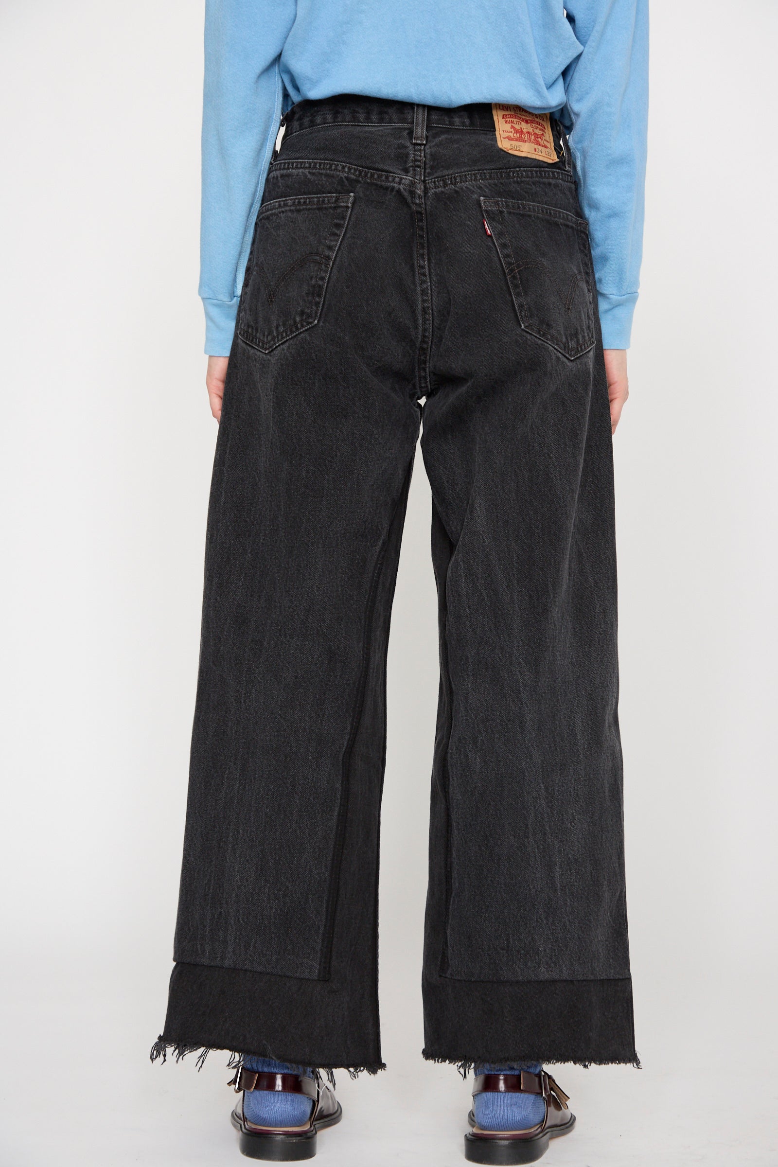 A person stands facing away, wearing a blue long-sleeve shirt and B Sides Reworked Culotte in Vintage Black with frayed hems.