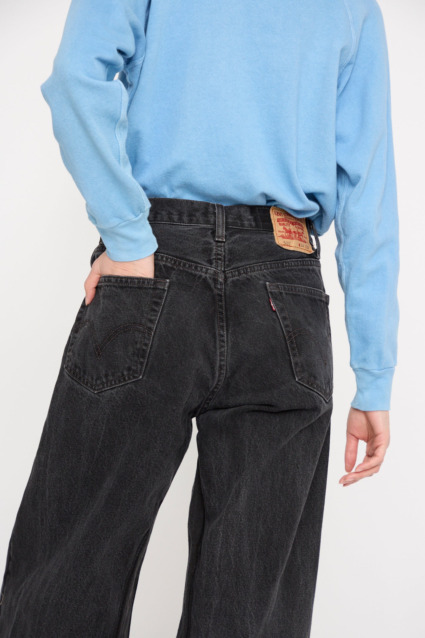 Wearing a blue shirt and black jeans with a visible leather patch, a model stands with one hand in their pocket, in the Reworked Culotte in Vintage Black by B Sides, against a white backdrop.