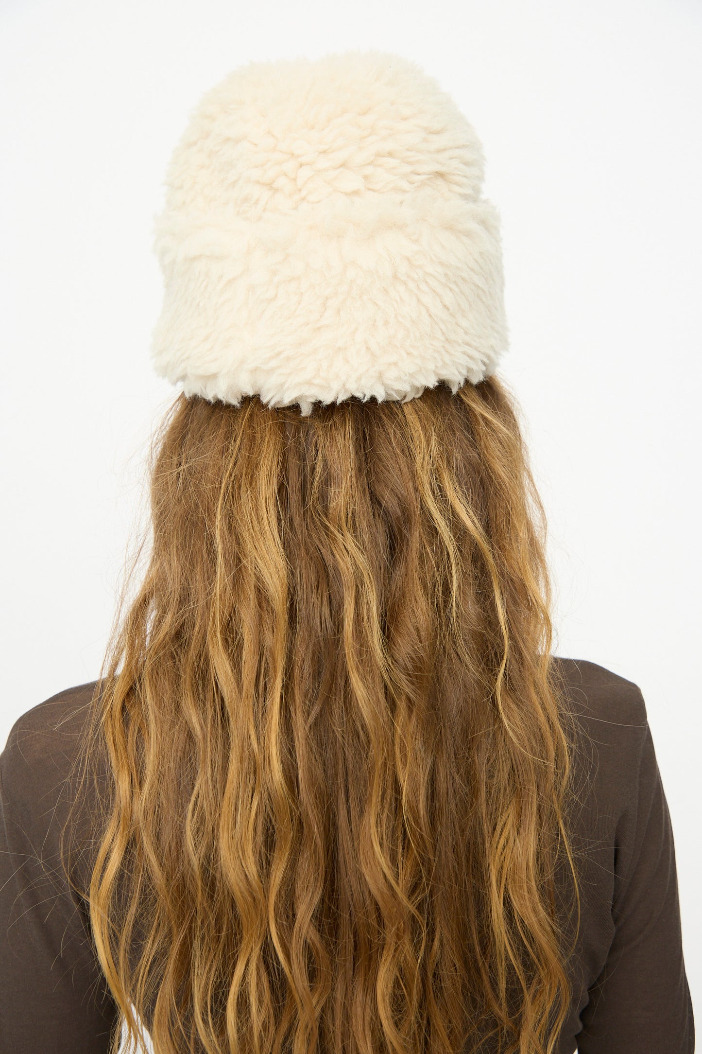 A person with long, wavy brown hair is shown from the back wearing the B Sides Sherpa Hat in Ivory, crafted from Japanese wool sherpa, and a brown top against a plain white background.