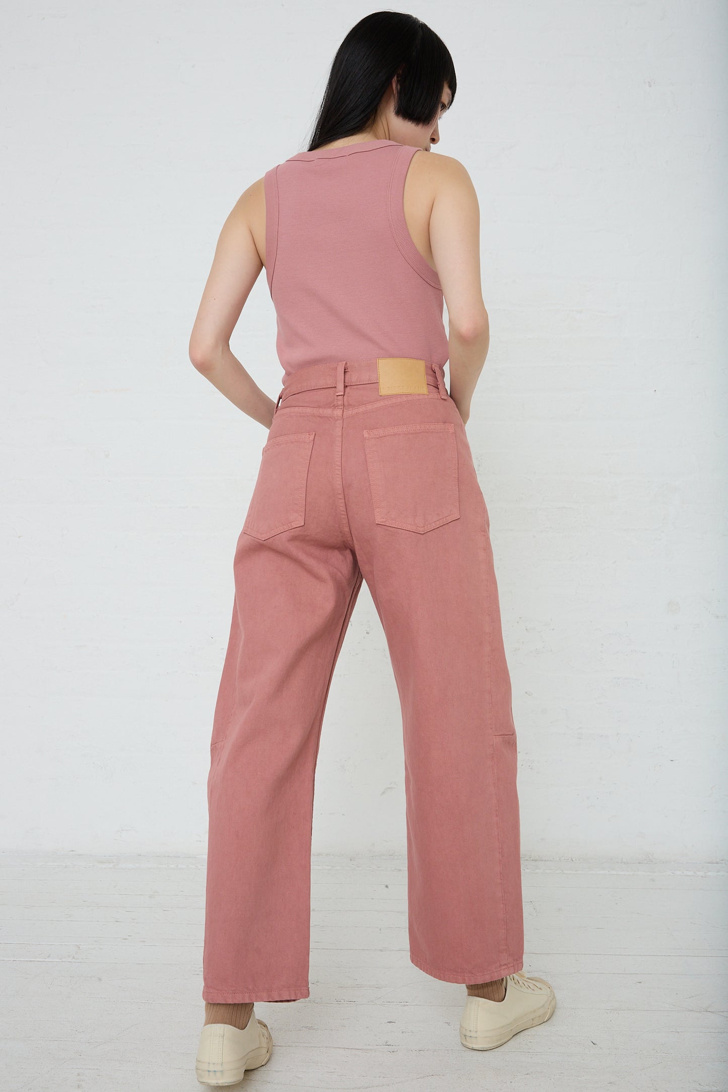 A woman seen from behind, wearing B Sides' Slim Lasso in Zinnia Overdye high-waisted denim trousers and a matching sleeveless top, standing in front of a white backdrop.