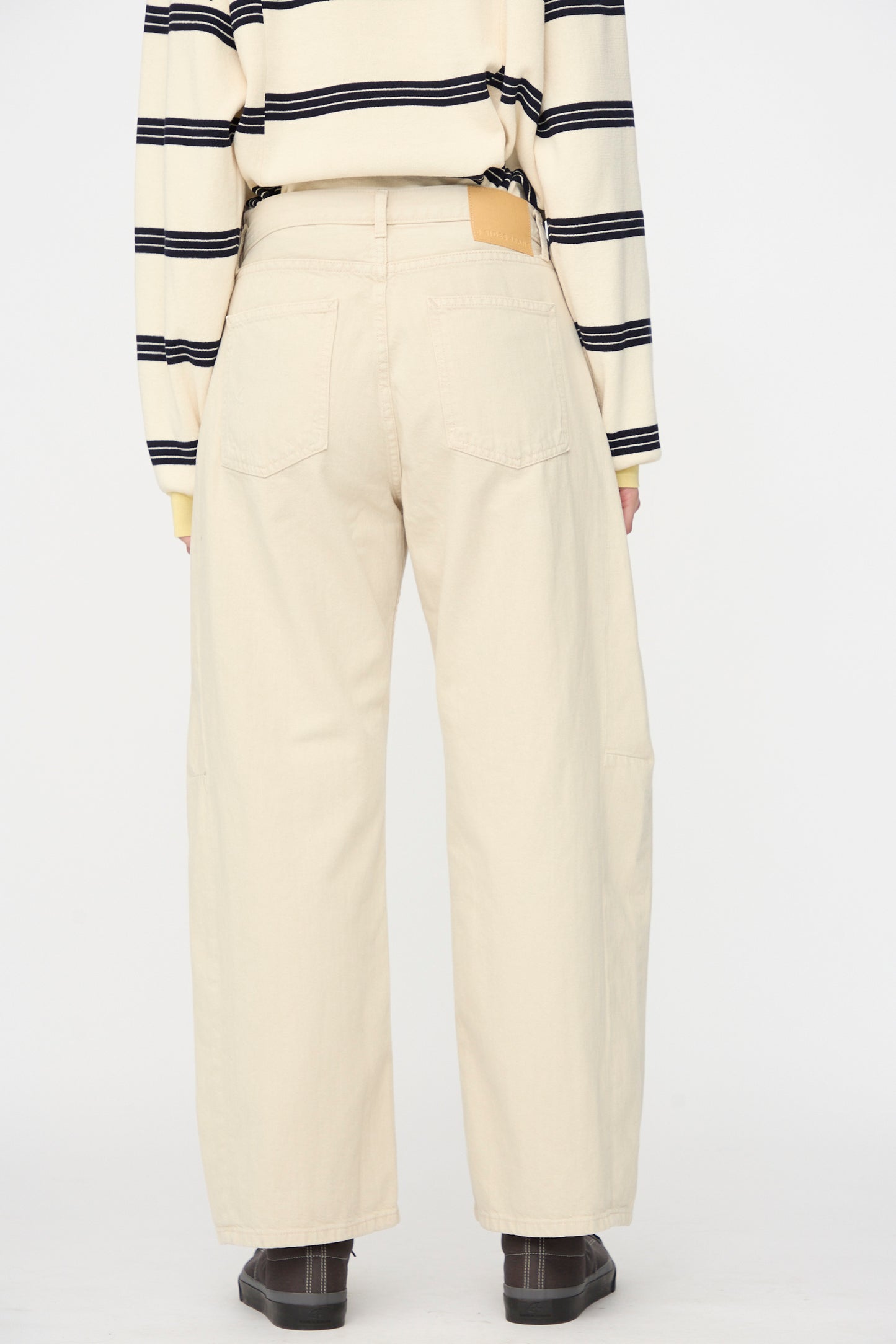A person stands with their back to the camera against a plain white background, wearing a beige long-sleeve striped shirt and B Sides Slim Lasso Jean in Chalk Overdye.