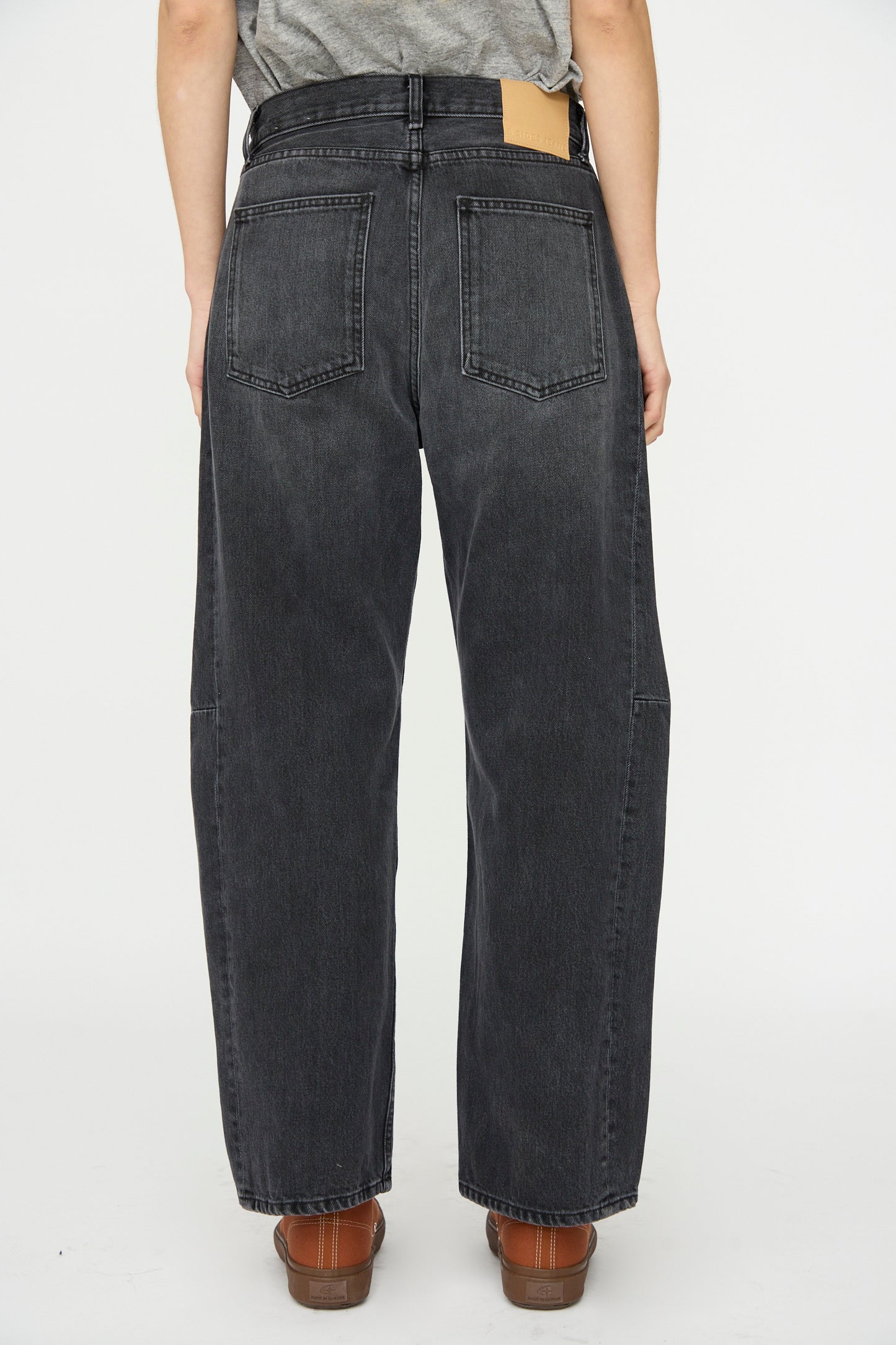 Person wearing B Sides Slim Lasso Jean in Stil Black, a high-waisted, slim-fit denim, paired with brown shoes, shown from the back against a plain white background.