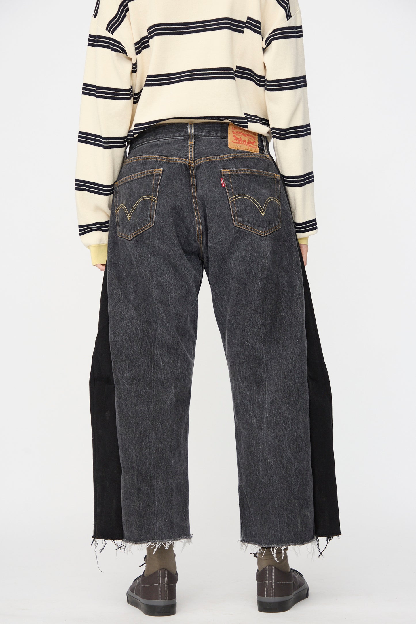 Person wearing a beige and black striped long-sleeve shirt, B Sides Vintage Lasso Jean in Black, and dark shoes, standing with their back to the camera.