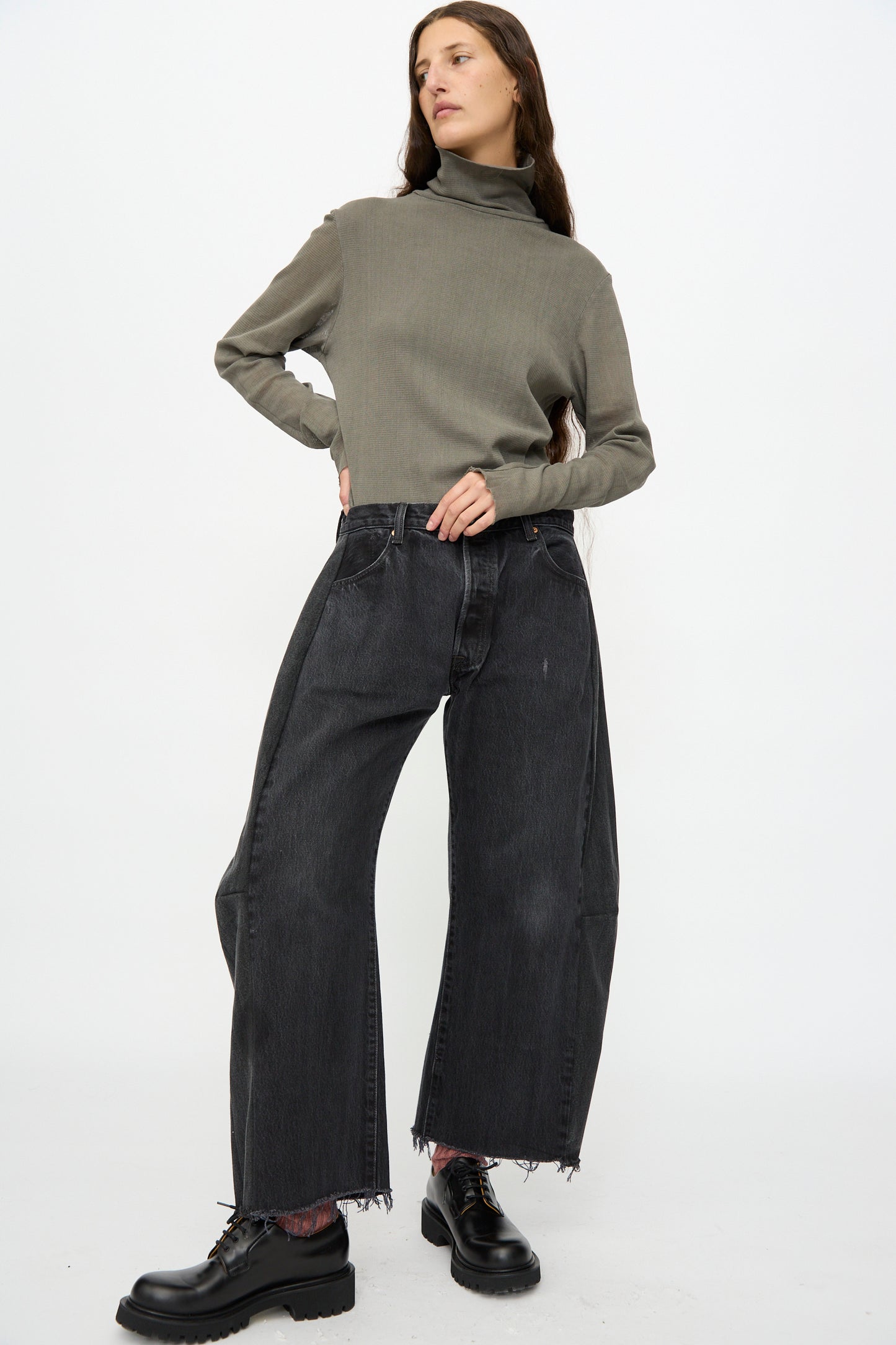 A person stands with one hand on hip, wearing a grey turtleneck sweater, B Sides Vintage Lasso Jean in Black, and black lace-up shoes against a plain white background.