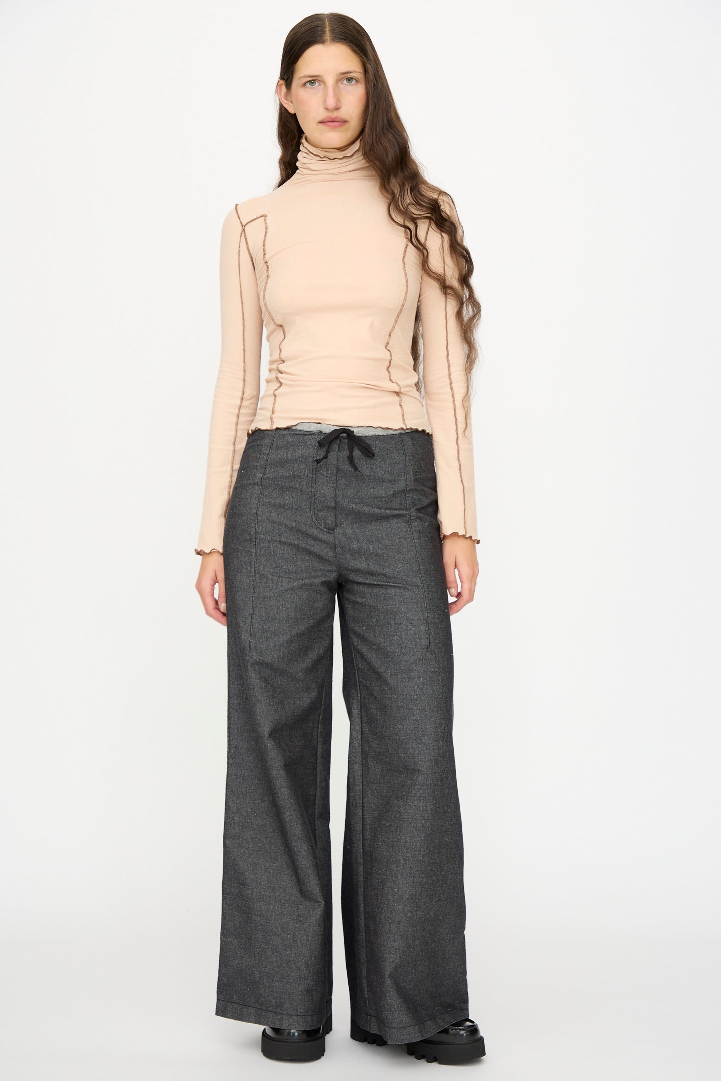 A person stands facing forward against a plain white background, wearing black Arthur Loose Pants by Baserange with a long-sleeve beige turtleneck top and black shoes.