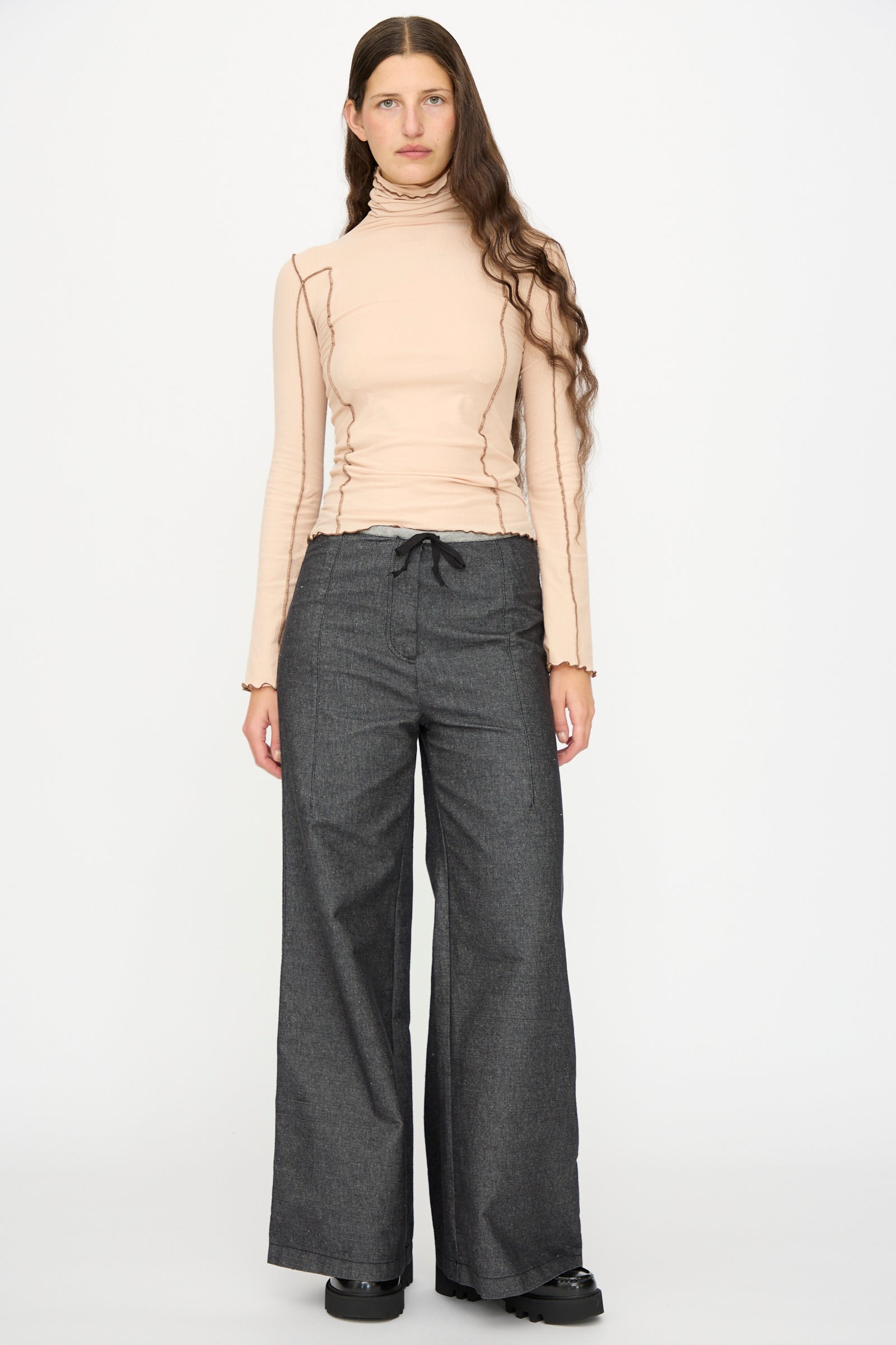 A person stands facing forward against a plain white background, wearing black Arthur Loose Pants by Baserange with a long-sleeve beige turtleneck top and black shoes.