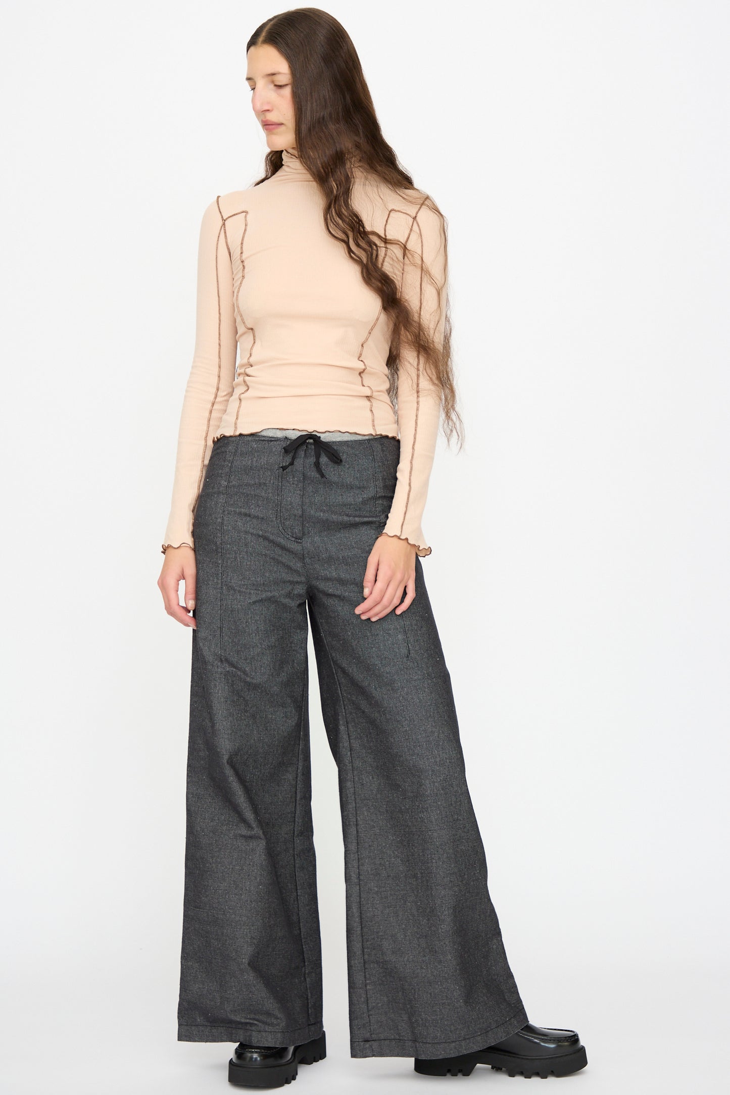 A person wears Baserange's Arthur Loose Pants in Black, paired with a beige long-sleeve top, standing against a plain white background and looking to the side.