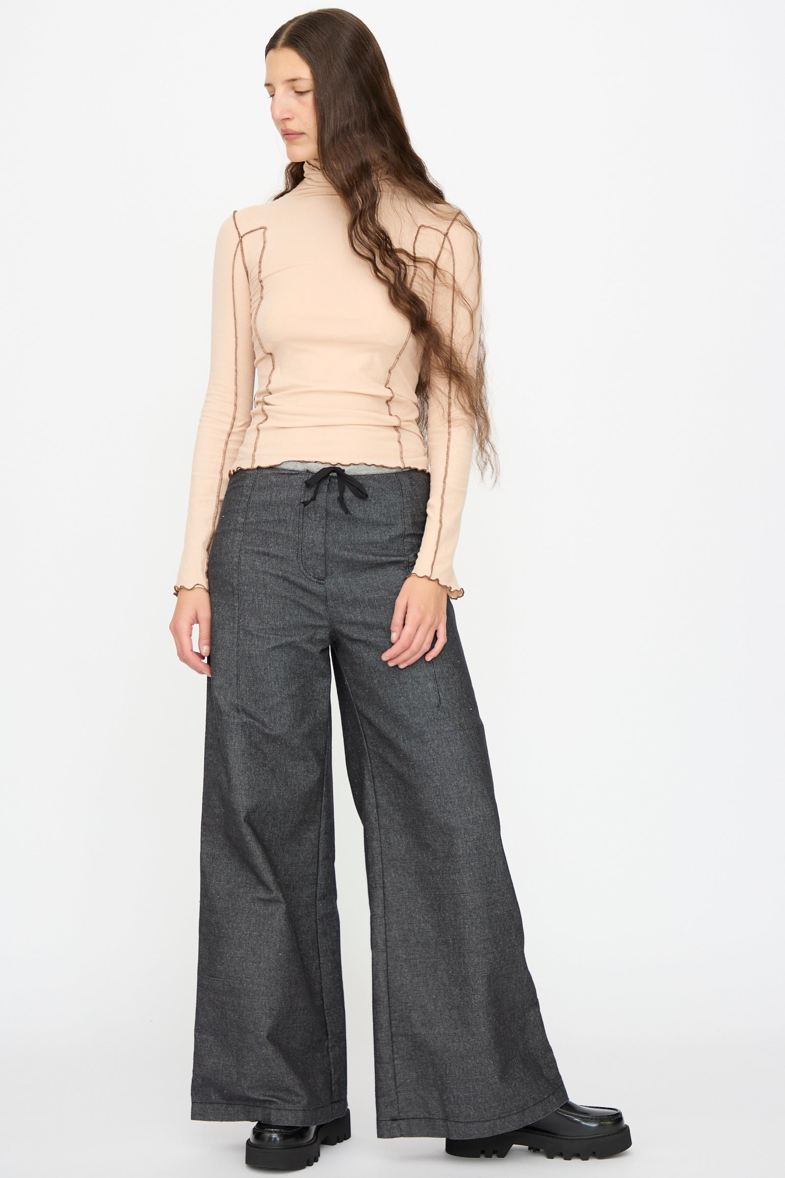 A person wears Baserange's Arthur Loose Pants in Black, paired with a beige long-sleeve top, standing against a plain white background and looking to the side.