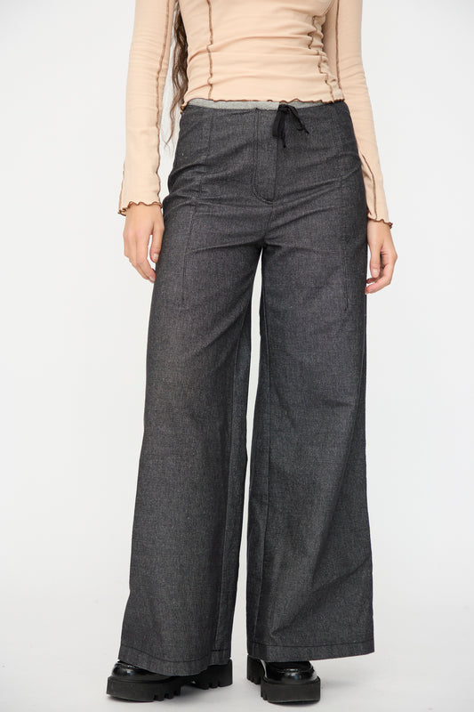 A person stands against a plain background wearing a beige long-sleeve top, black shoes, and Baserange Arthur Loose Pants in Black.