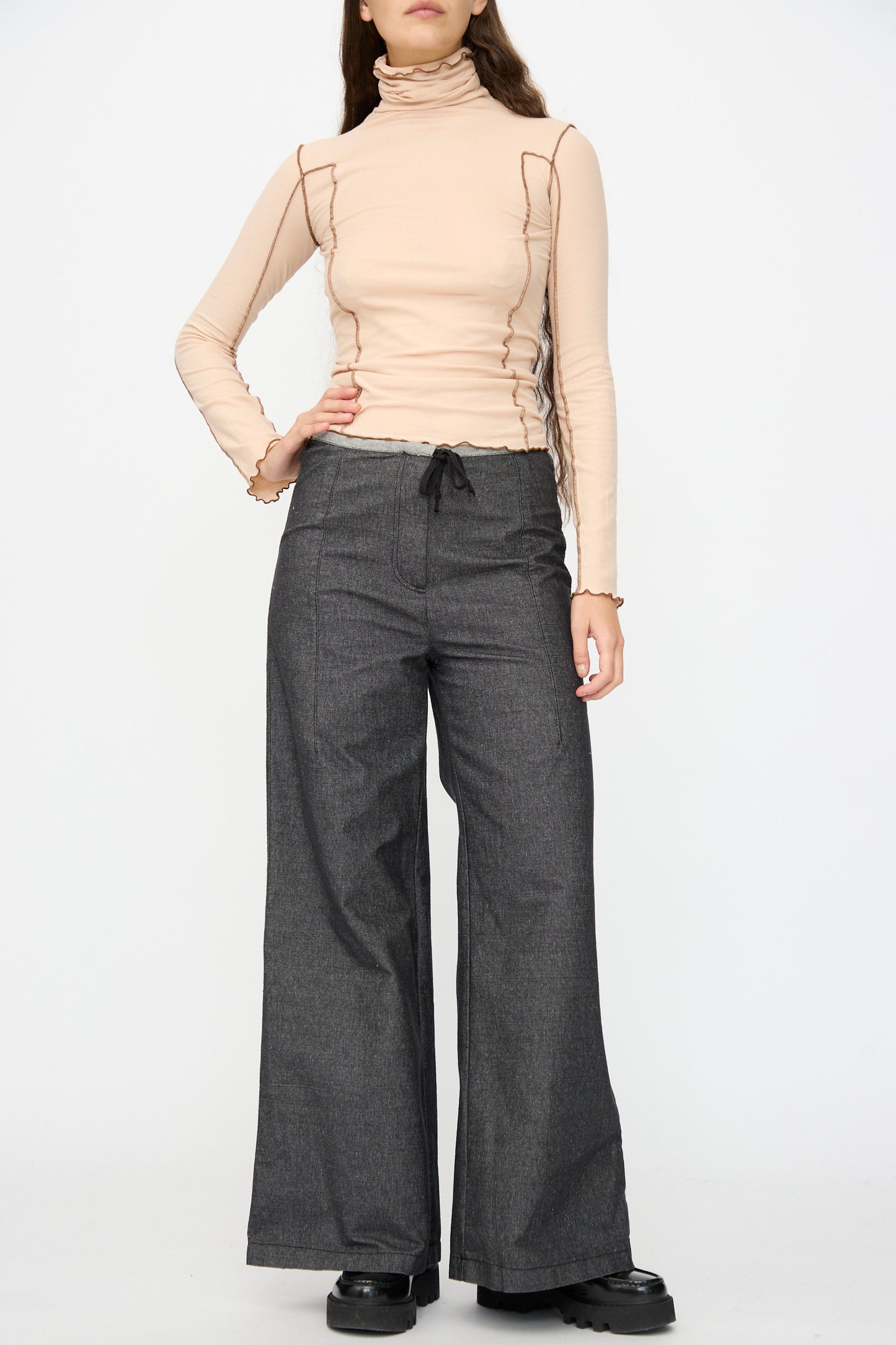 A person stands against a plain white background wearing a beige long-sleeve turtleneck top, black Baserange Arthur Loose Pants, and black platform shoes.
