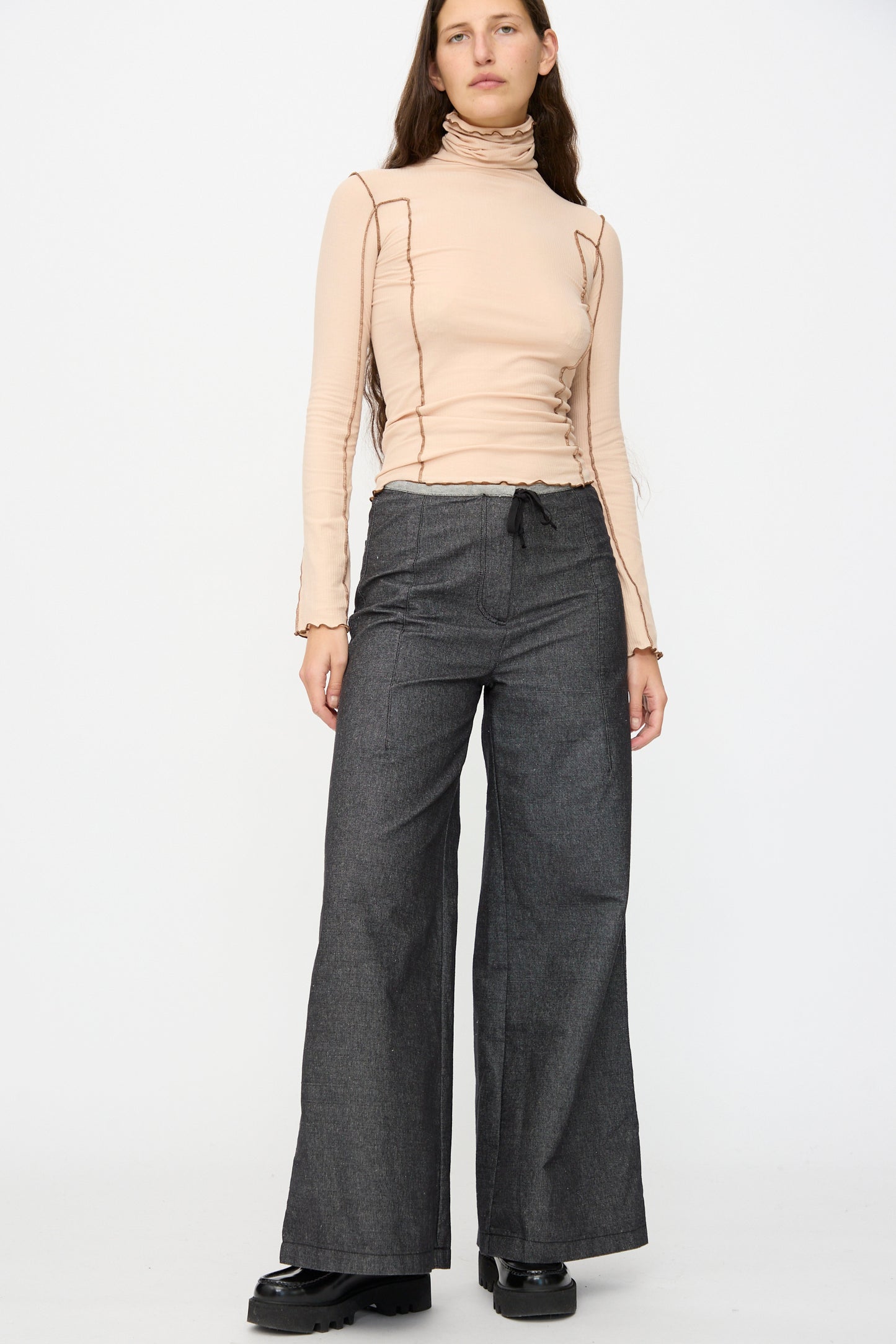 A person stands against a plain background wearing a beige turtleneck long-sleeve top with exposed seams, paired with Baserange Arthur Loose Pants in Black and black shoes.