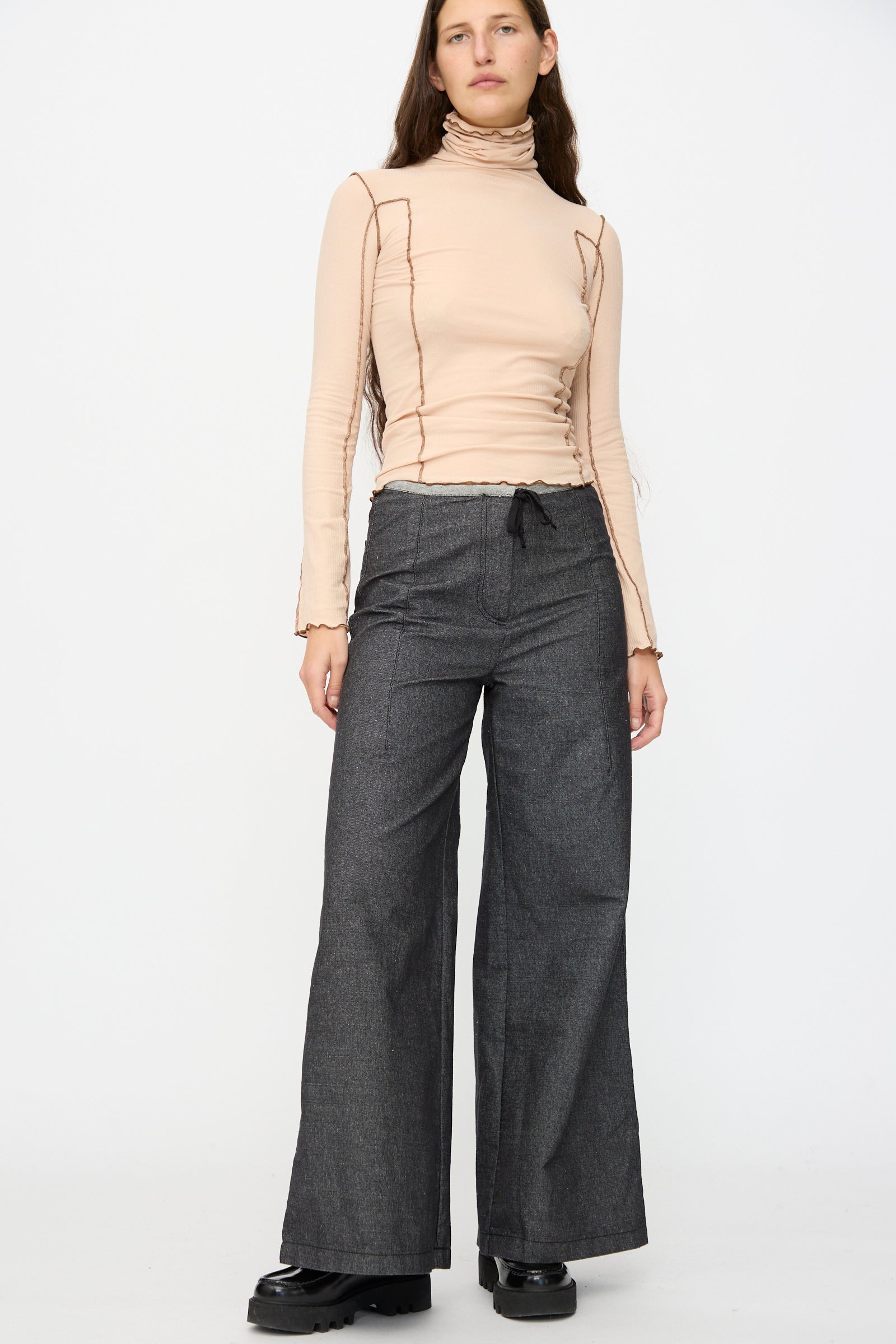 A person stands against a plain background wearing a beige turtleneck long-sleeve top with exposed seams, paired with Baserange Arthur Loose Pants in Black and black shoes.