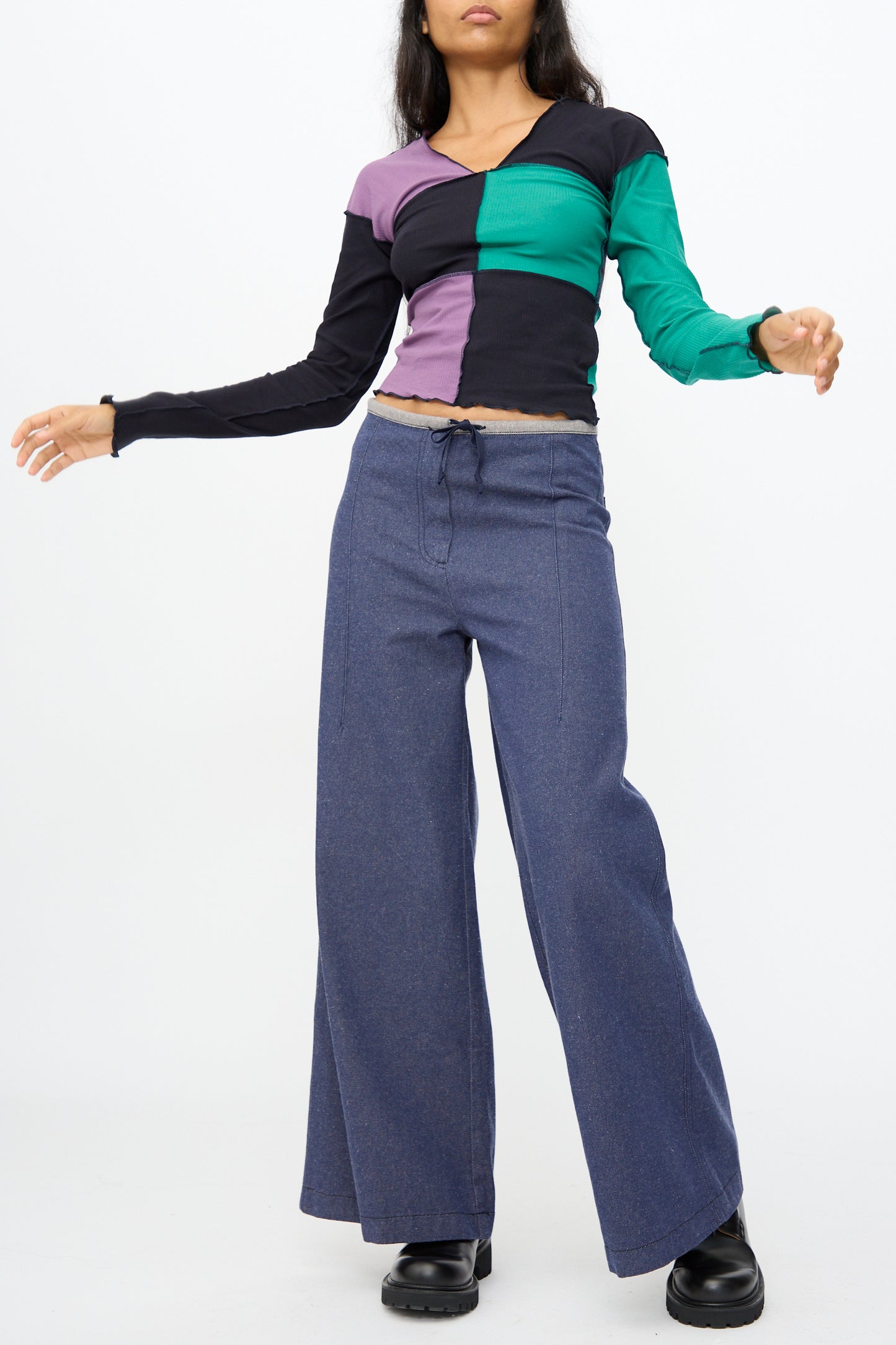A person in a green, purple, and black long-sleeve colorblock top, Baserange Arthur Loose Pants in Recycled Indigo, and black shoes stands against a plain white background.