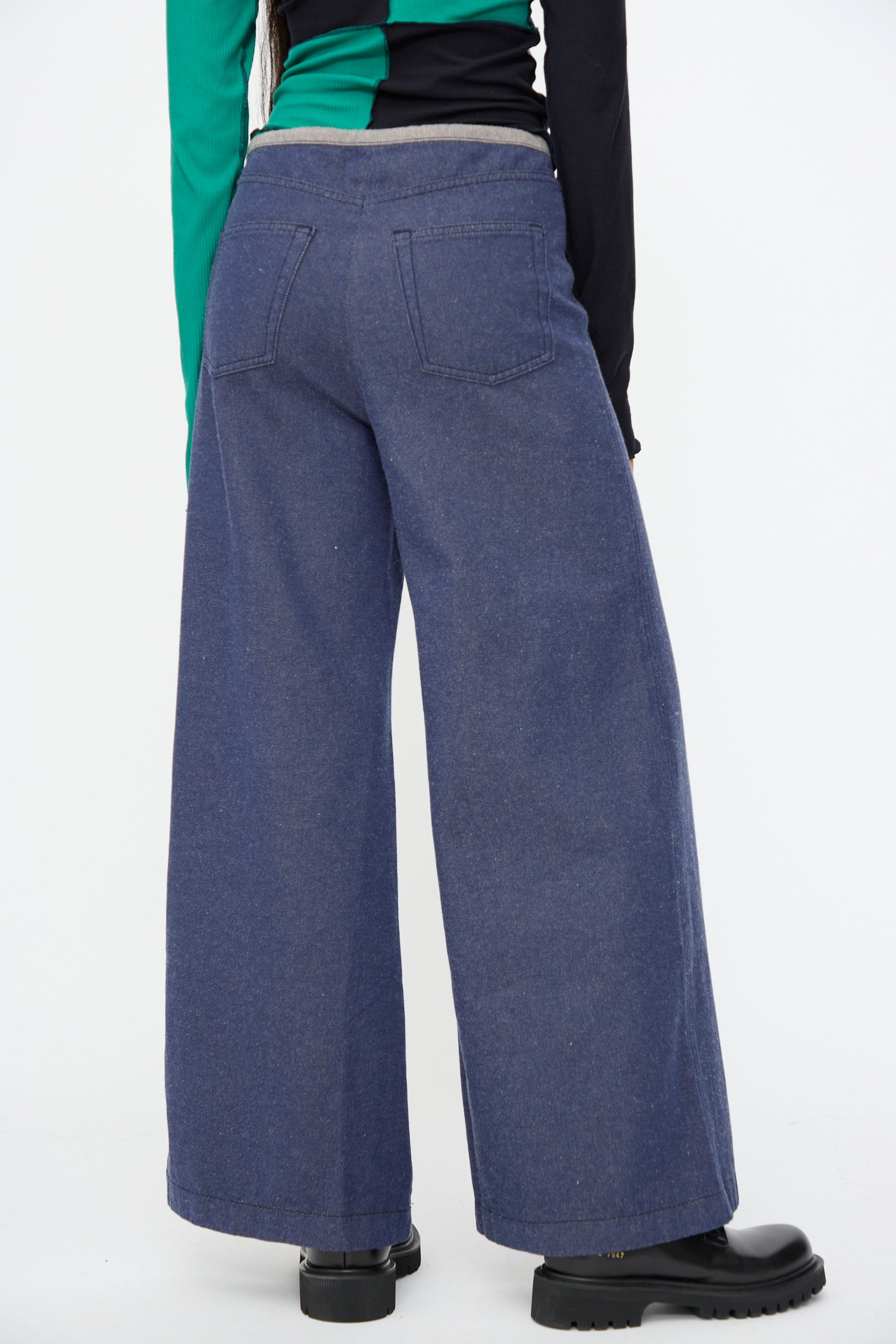 Model wears Baserange Arthur Loose Pants in Recycled Indigo, styled with black boots and a long-sleeve color-blocked shirt, standing against a plain white background.