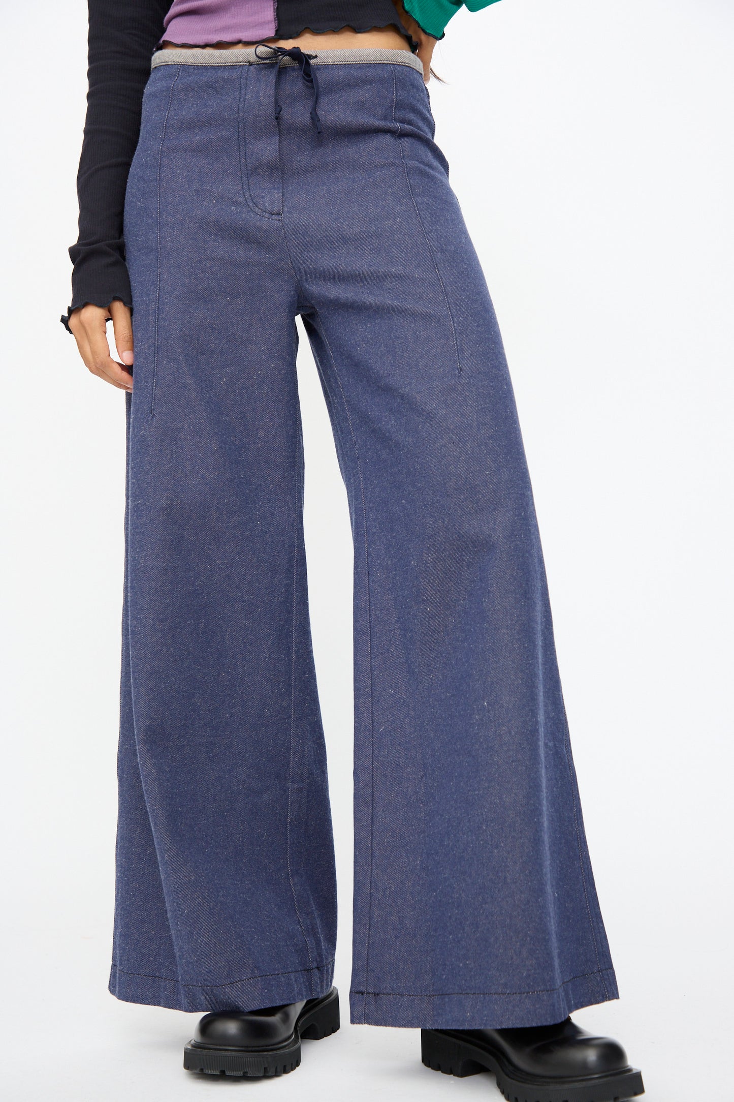 Person wearing Baserange Arthur Loose Pants in Recycled Indigo—wide-leg, flared denim—paired with black platform shoes and a partial view of a color-blocked long sleeve top.