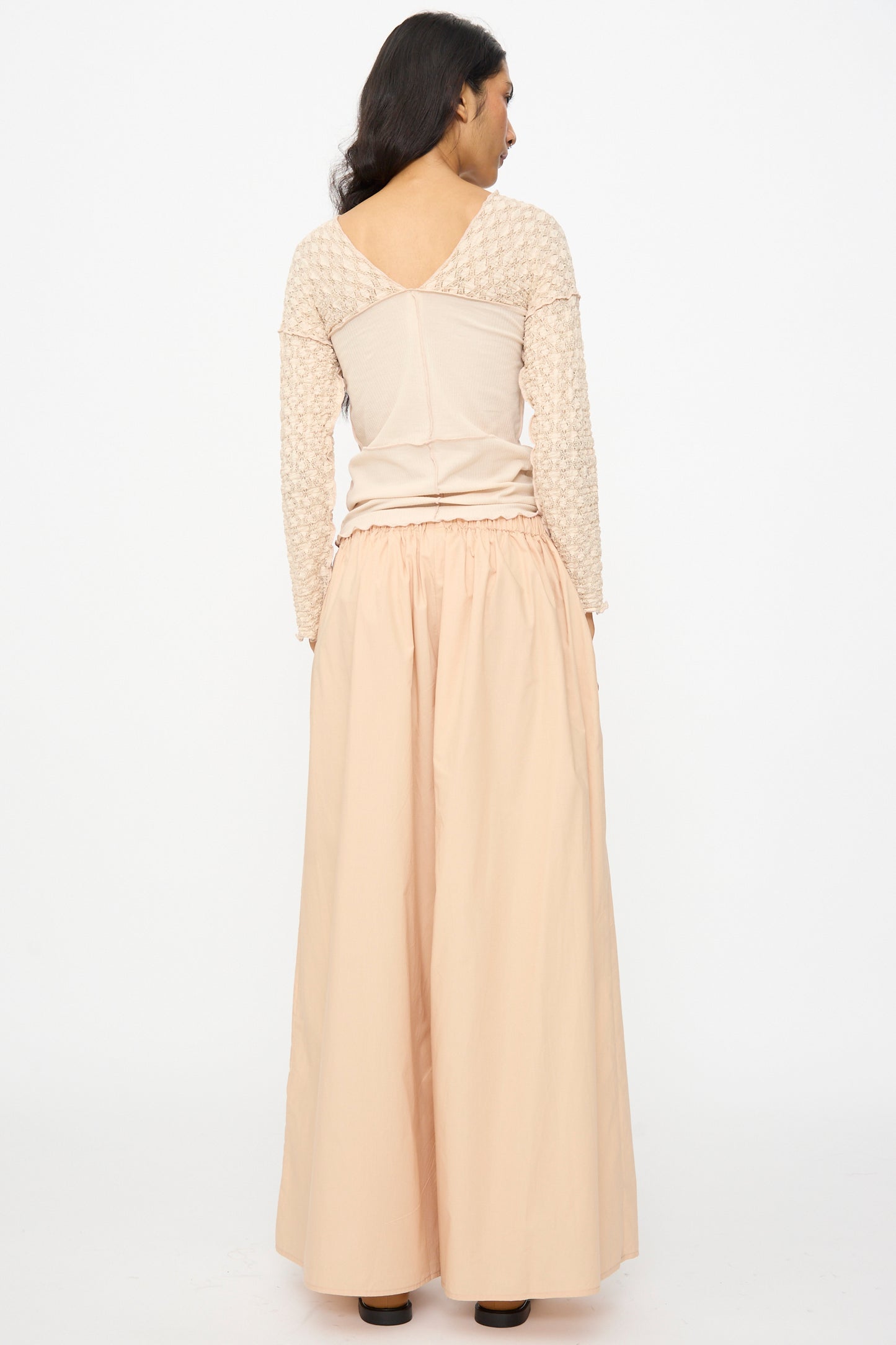 A woman stands facing away, wearing a beige long-sleeve top and Baserange Cable Pants in Andradita Sand, featuring a subtle pattern, light peach color, wide cotton legs, and an elastic waist.