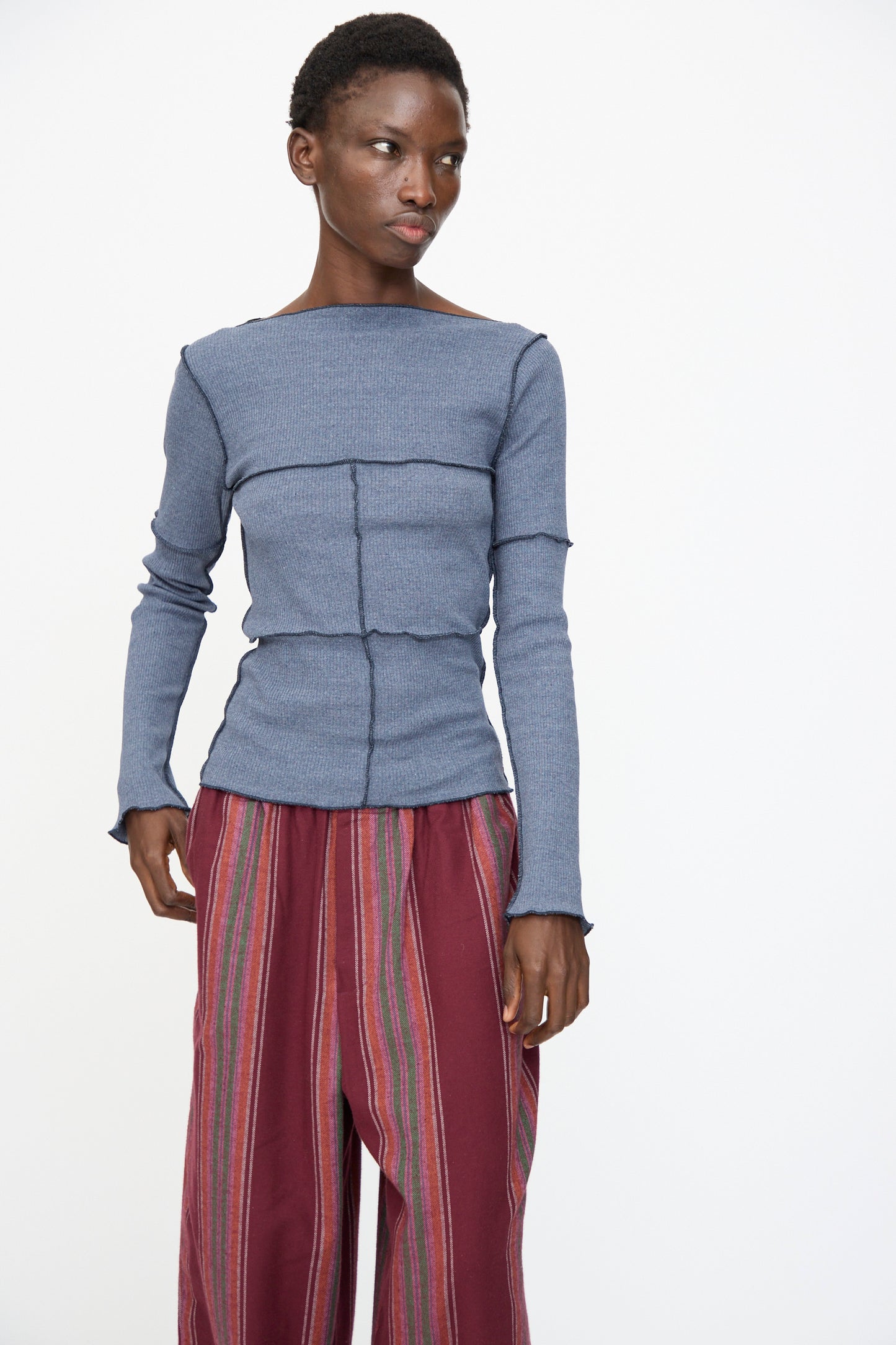 Model wears the Baserange Cinder Long Sleeve Tee in Dark Denim with loose maroon striped pants, standing against a plain white background.