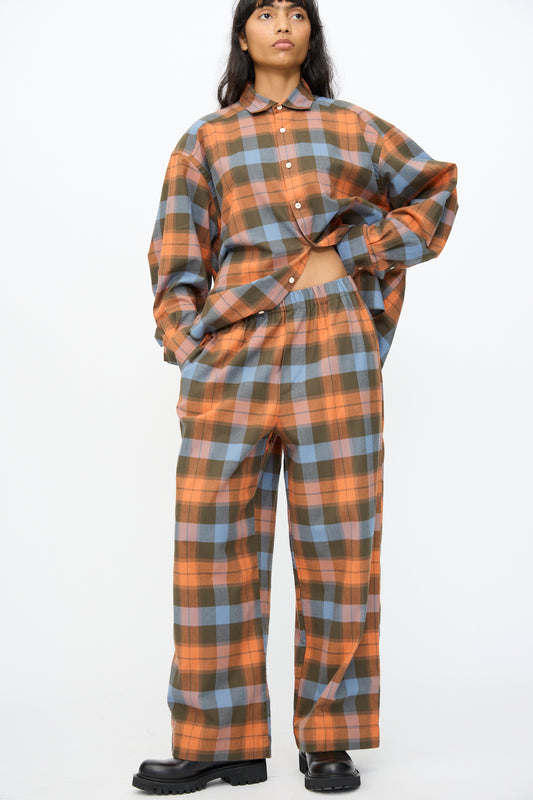 A person wearing Baserange's Claude Pant in Frame Plaid, styled with a matching button-up shirt in orange, blue, and gray tones and black boots, stands against a plain white background.