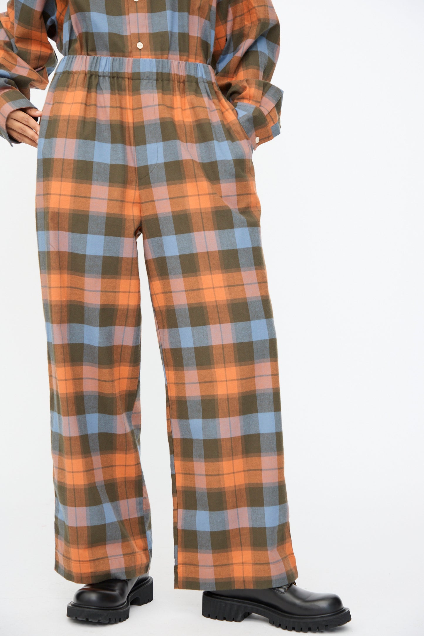 A person wearing the Baserange Claude Pant in Frame Plaid—orange, olive green, and blue organic cotton wide-leg pants—cropped at the waist with black chunky shoes.